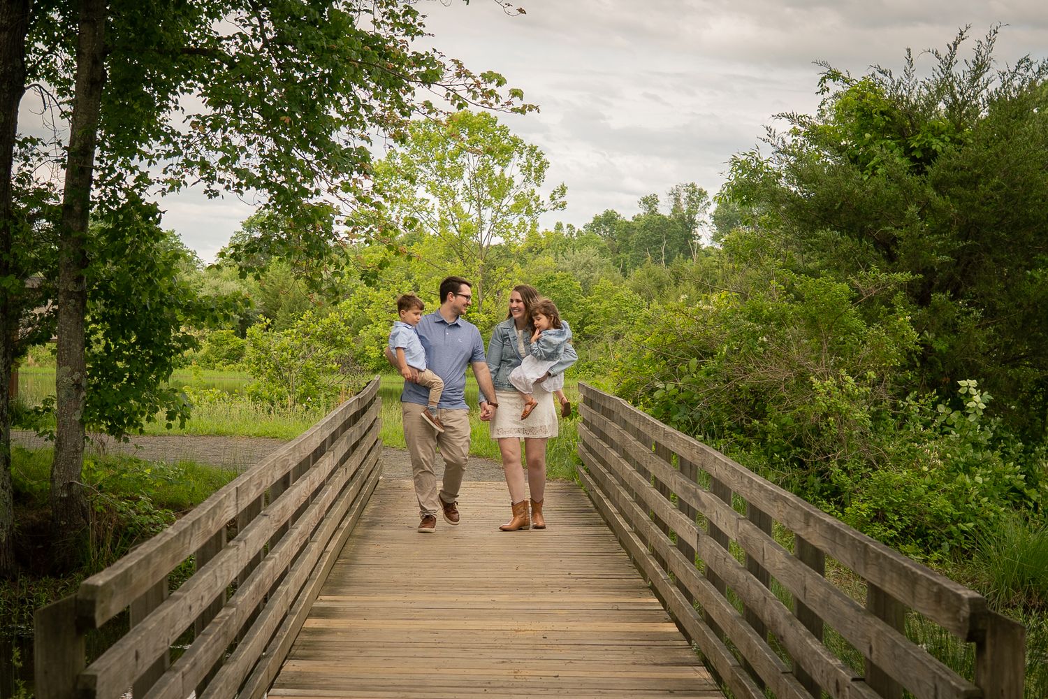 A joyful family walks together along a wooden bridge pathway surrounded by lush summer greenery.