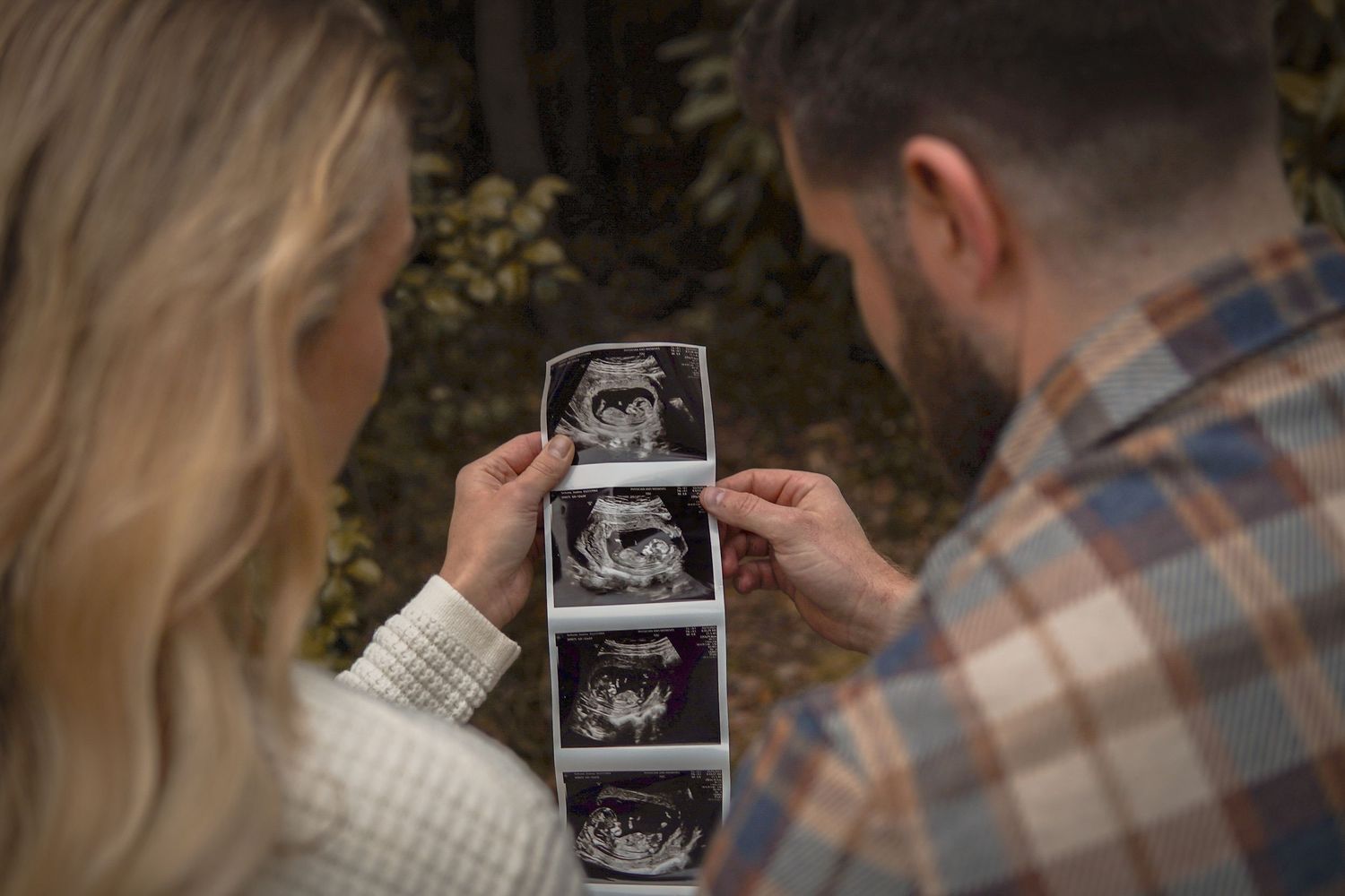 A couple holds and examines ultrasound sonogram photos together outdoors in a natural setting.
