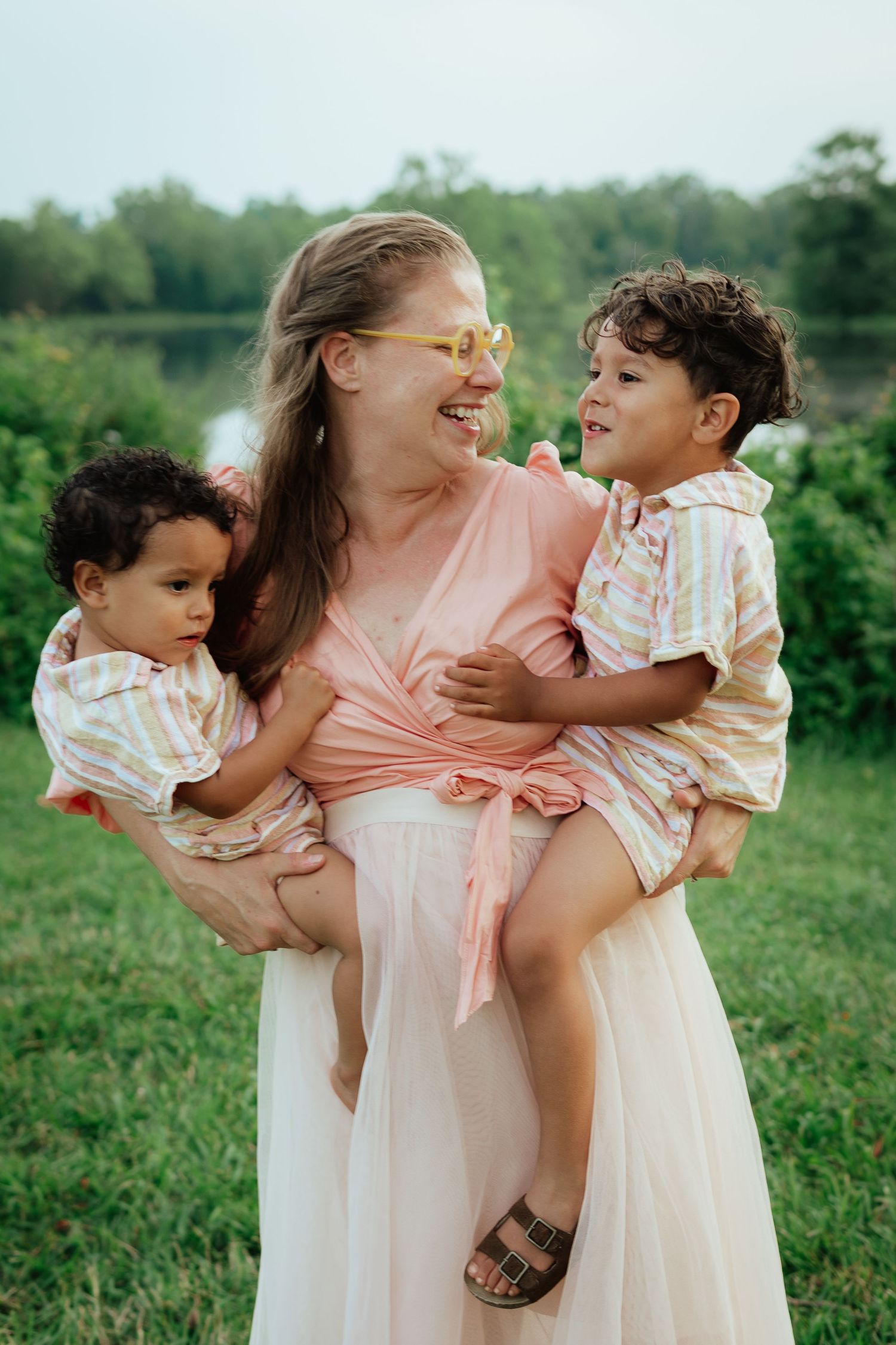 Parent wearing pink dress holds two children while laughing outdoors.