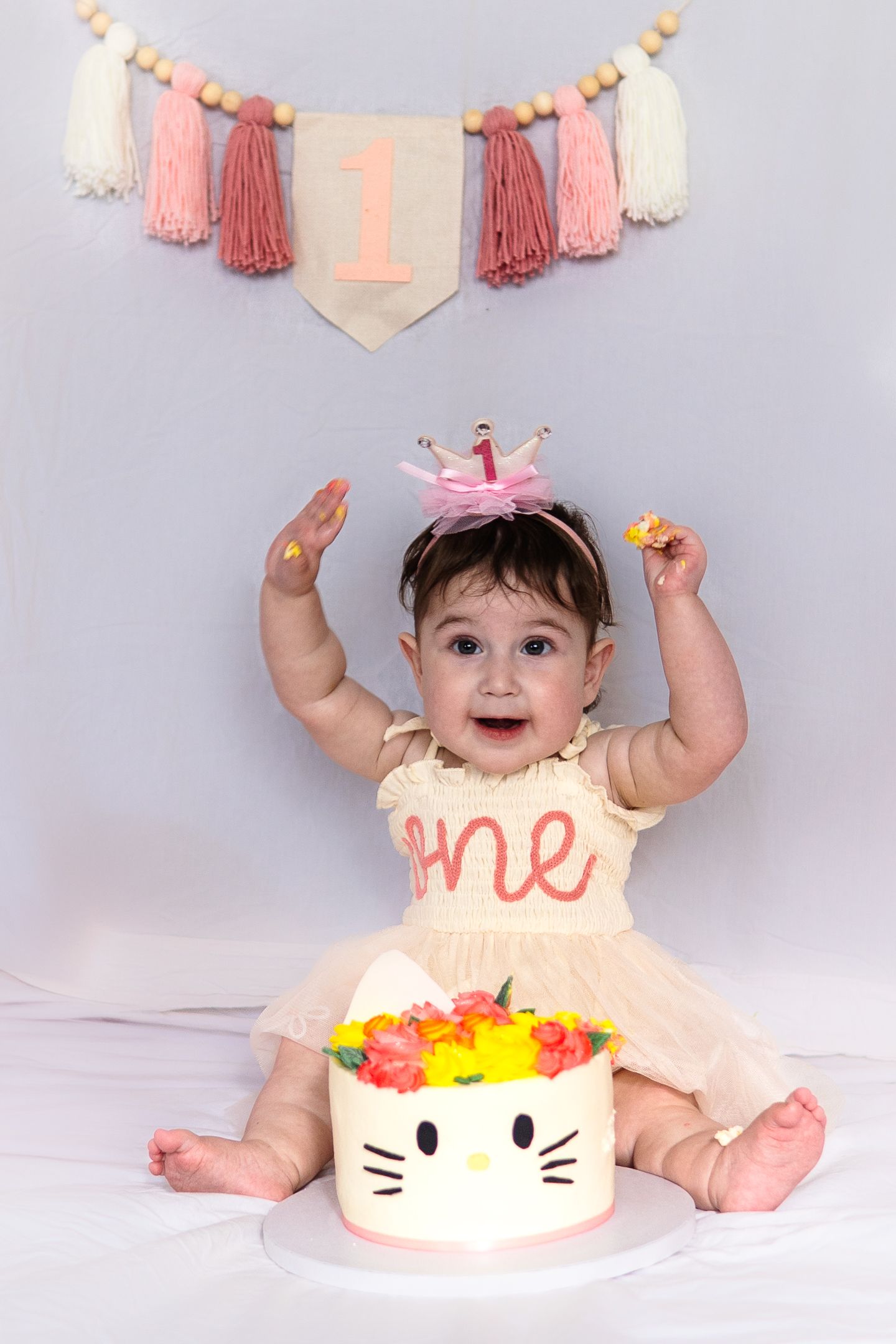 First birthday celebration with a Hello Kitty themed cake, pink tasseled banner, and peach dress decor against gray backdrop.