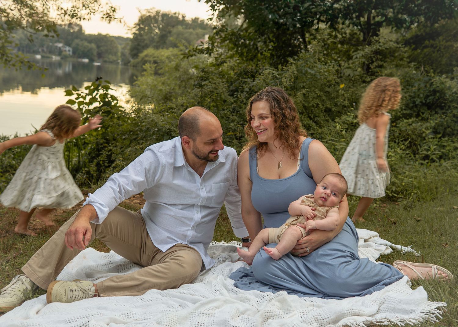 Family sitting on white blanket by lake at sunset, parents with baby and two children playing around them.