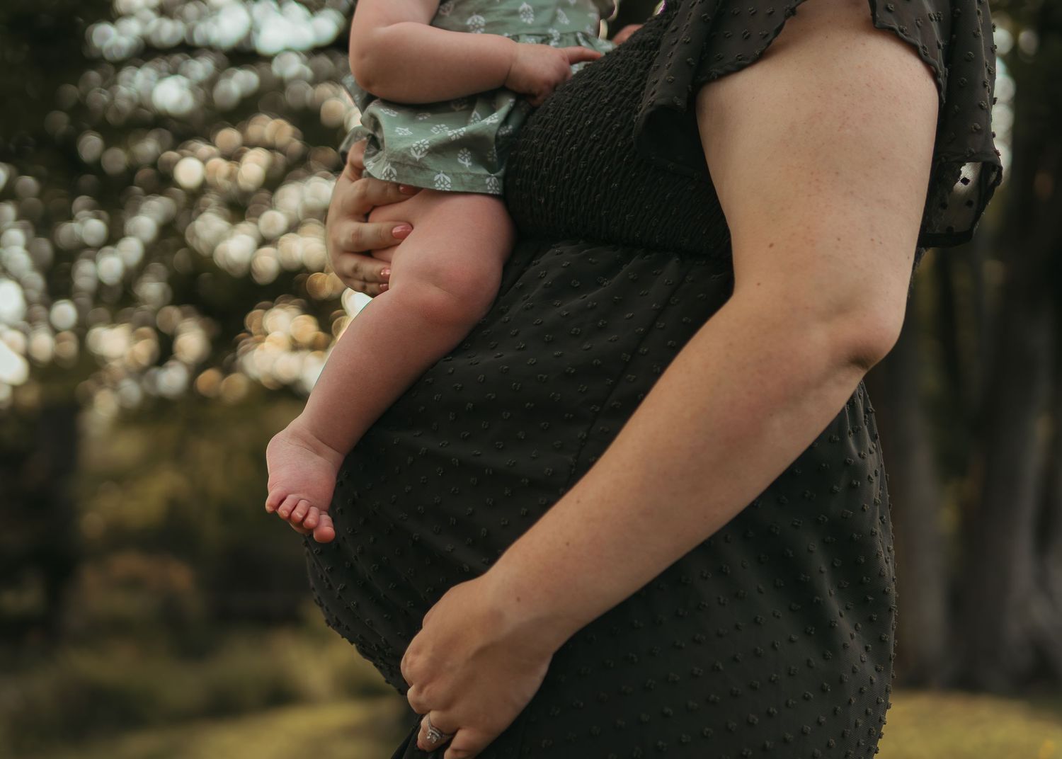 A mother in a dark dress holds her newborn baby outdoors, with soft bokeh trees in the background.
