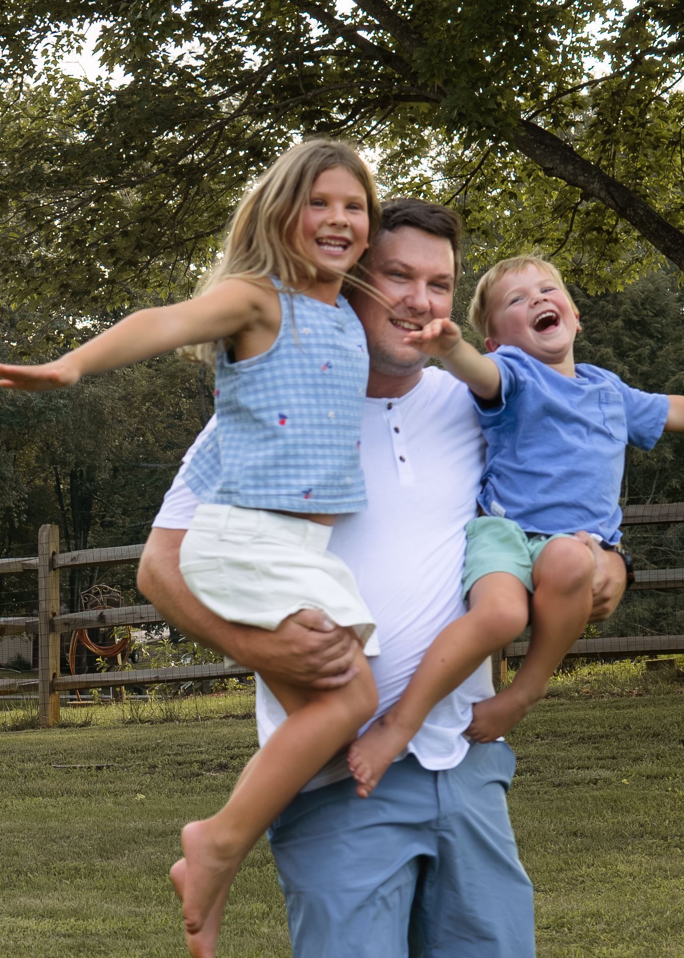 A laughing father carries two joyful children on his arms outdoors in a sunny green backyard.