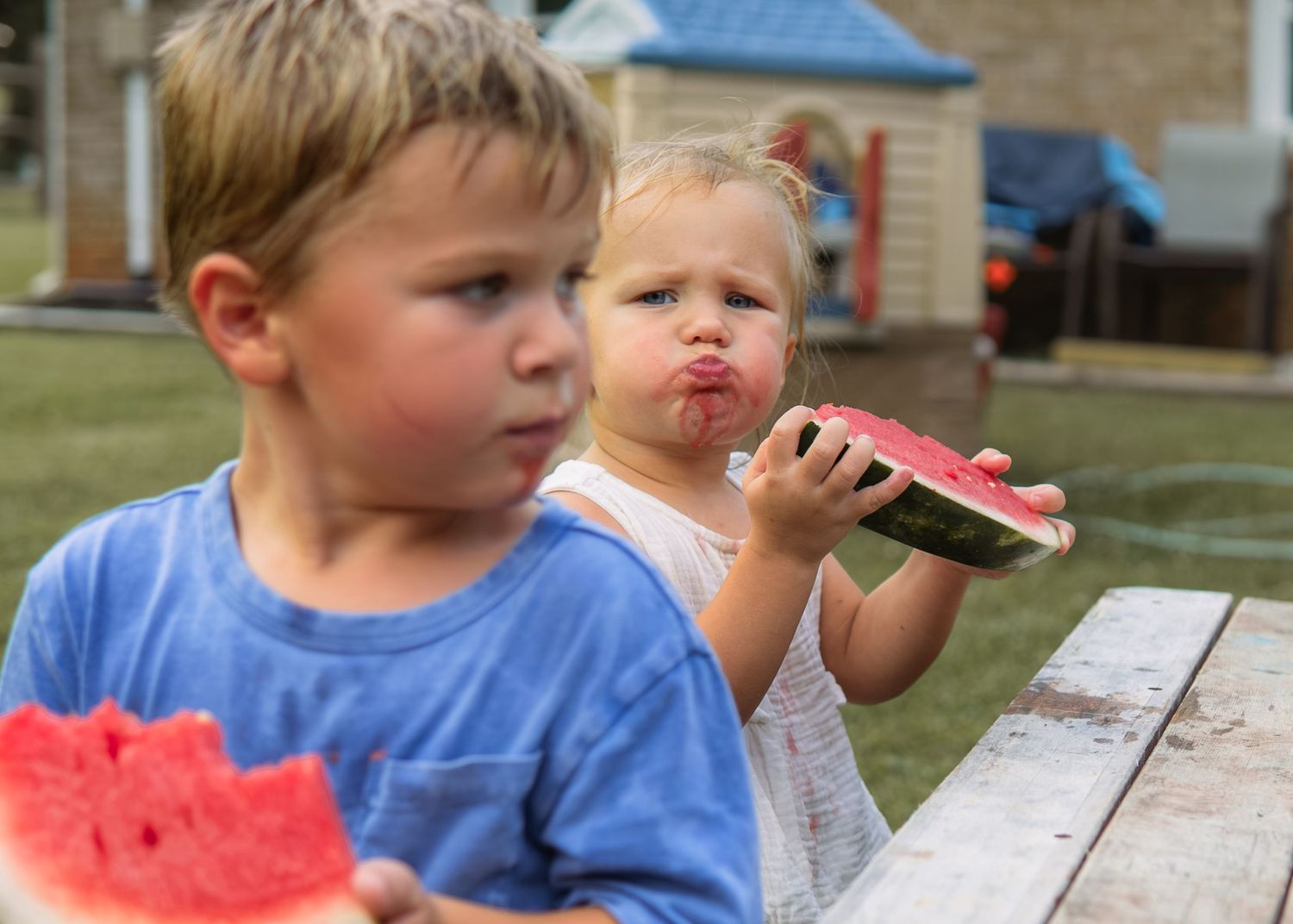 Two young children eating fresh watermelon slices at an outdoor picnic table on a summer day.