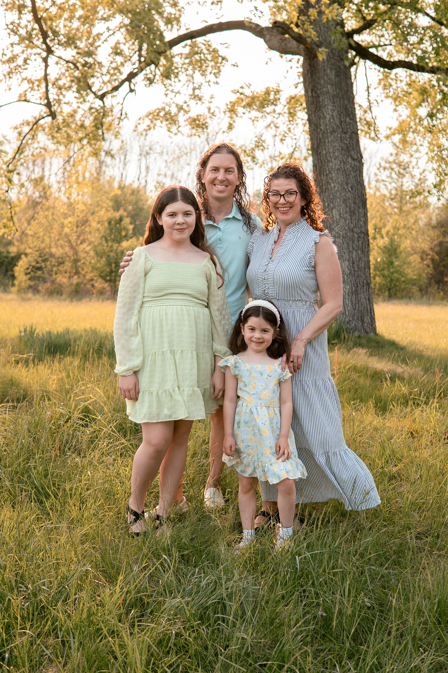 A family poses together in a sunlit meadow beneath a large tree during golden hour, wearing light-colored dresses.