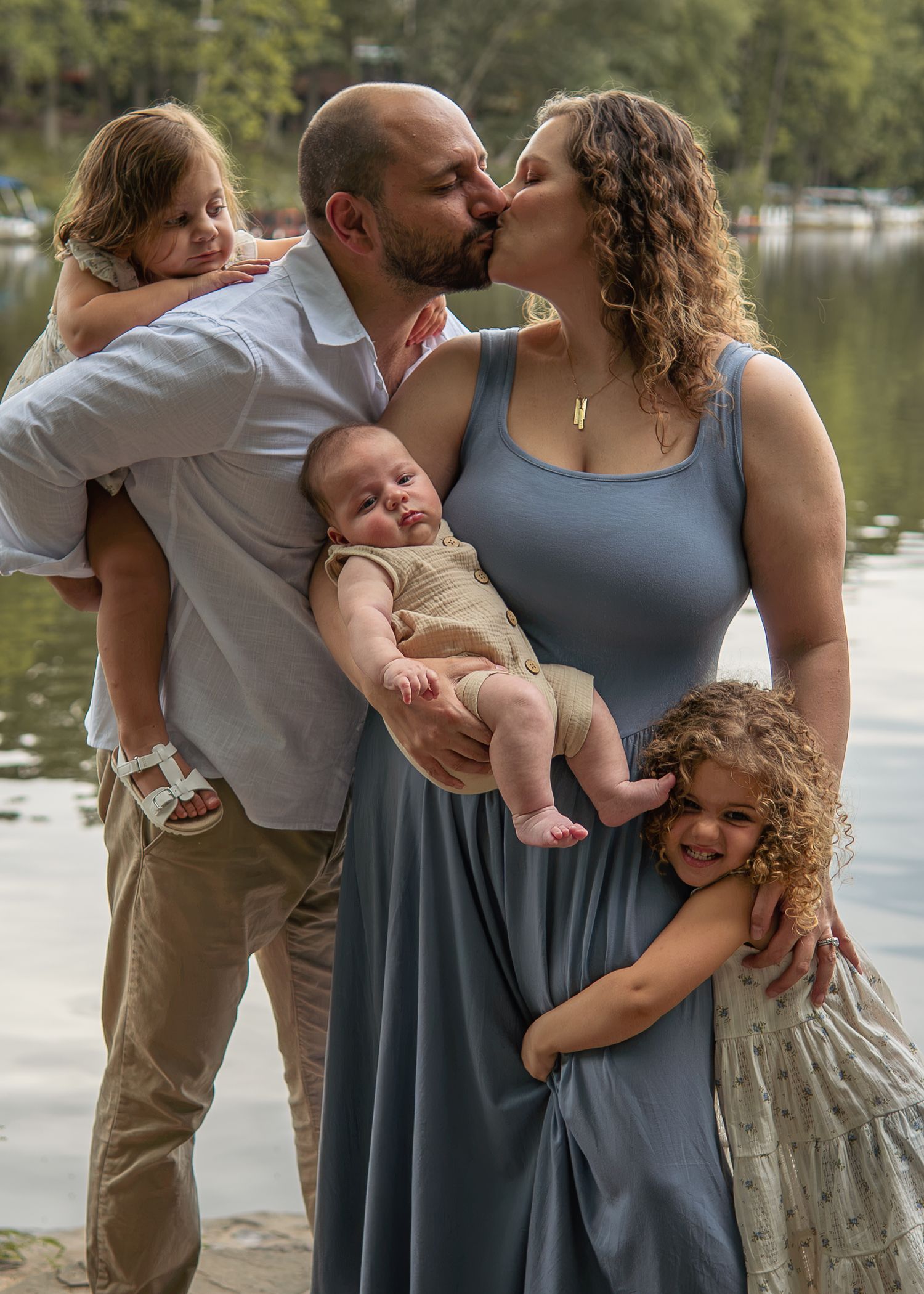 Family of five posing by a lake, parents kissing while holding baby and two young children embracing them.