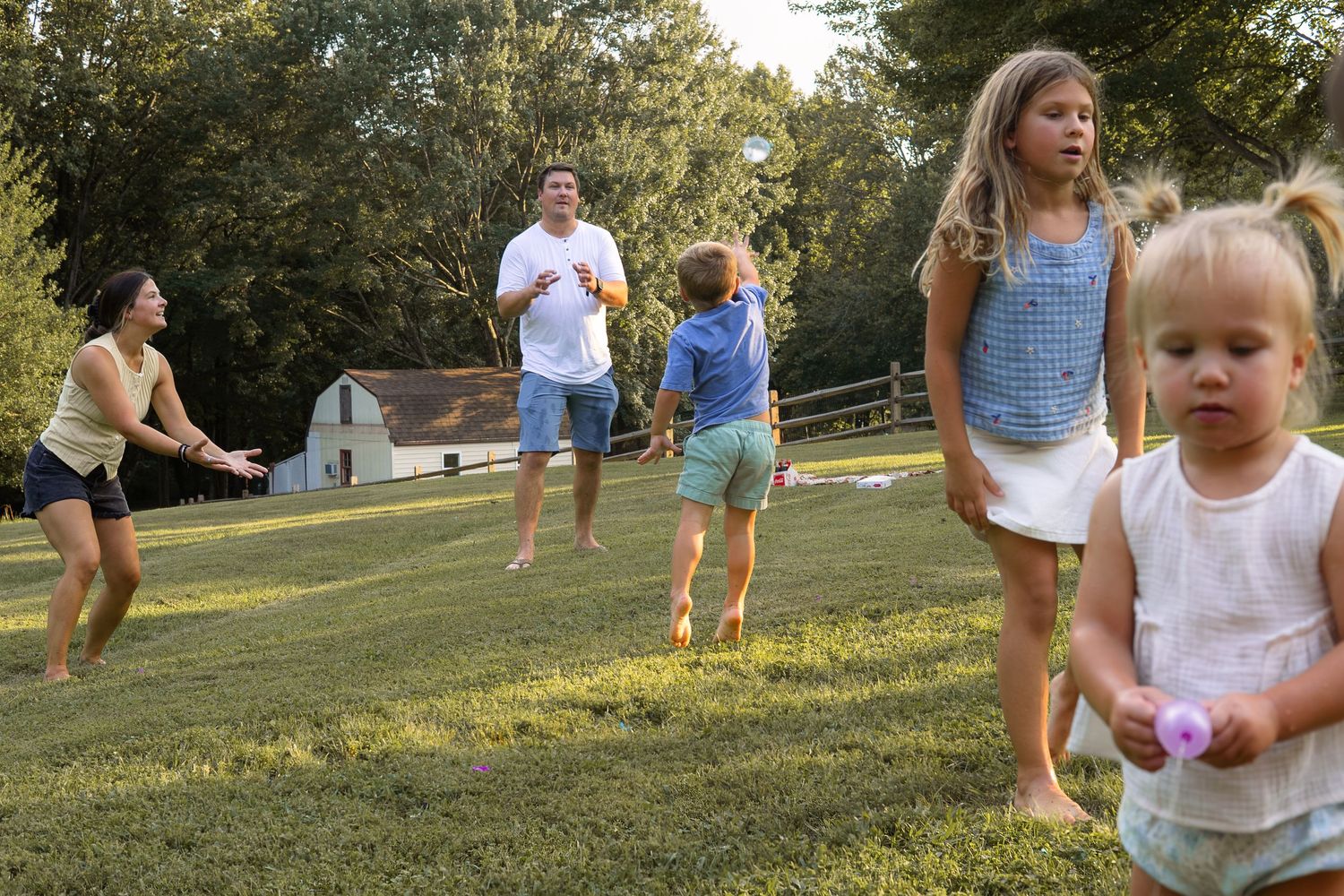 Family playing catch together in a sunny backyard with a barn in the background.