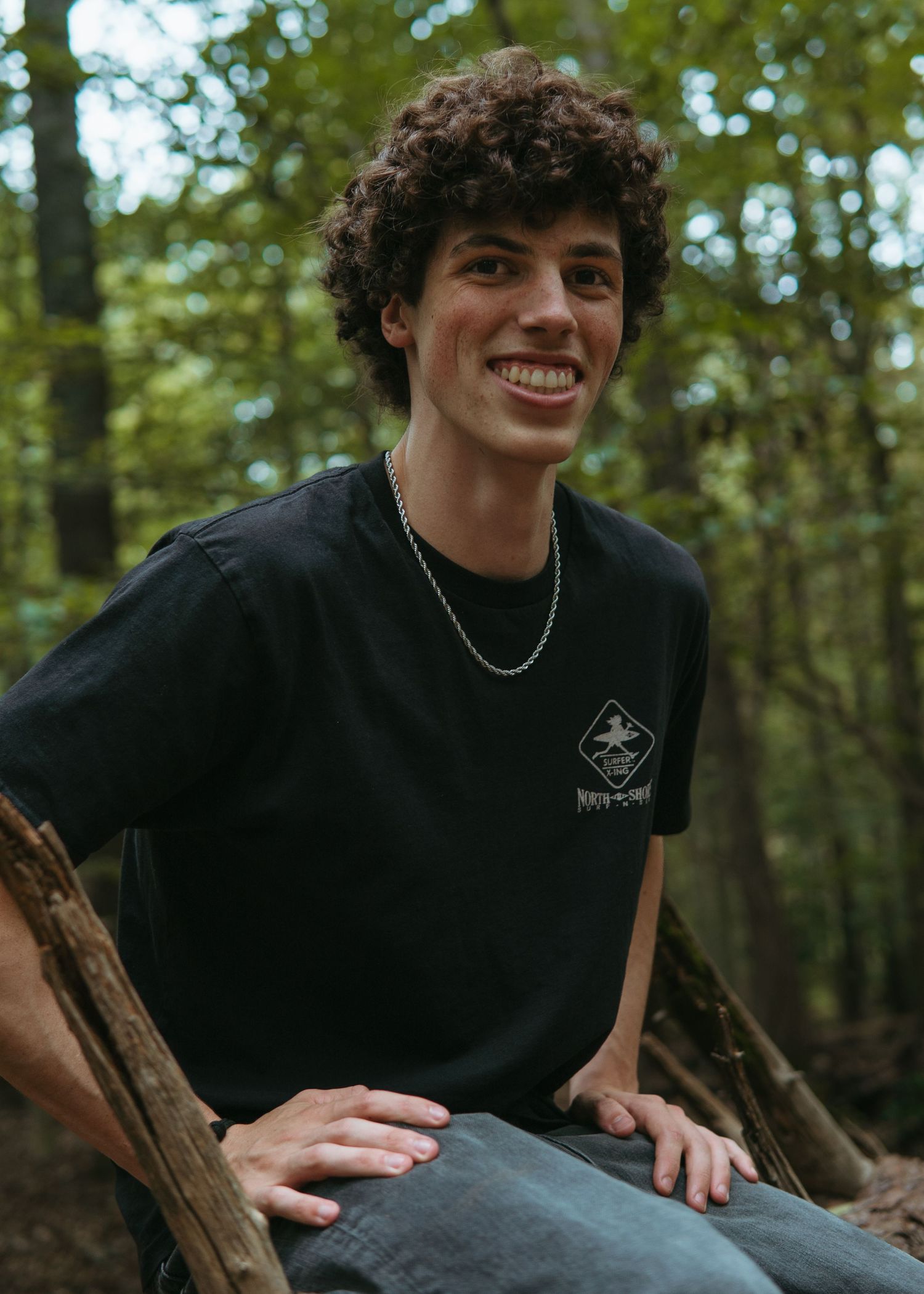 Smiling young man with curly hair wearing a black graphic t-shirt and silver chain necklace, sitting outdoors in a forest.