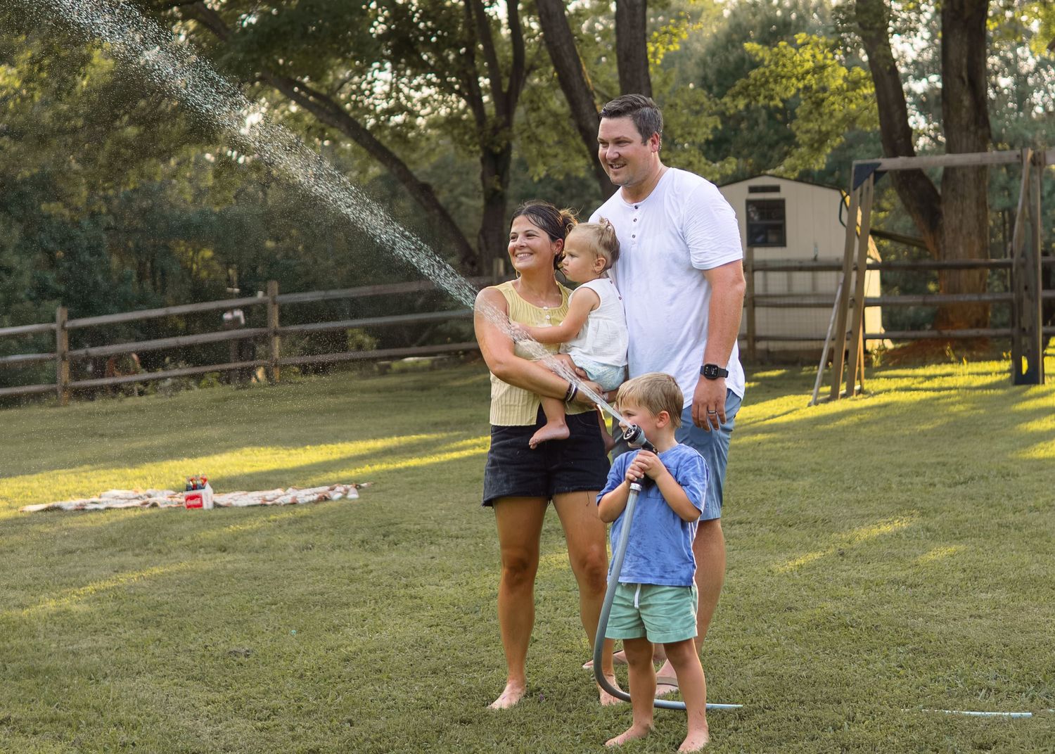A happy family of four plays in sprinkler water on a sunny green lawn outdoors.