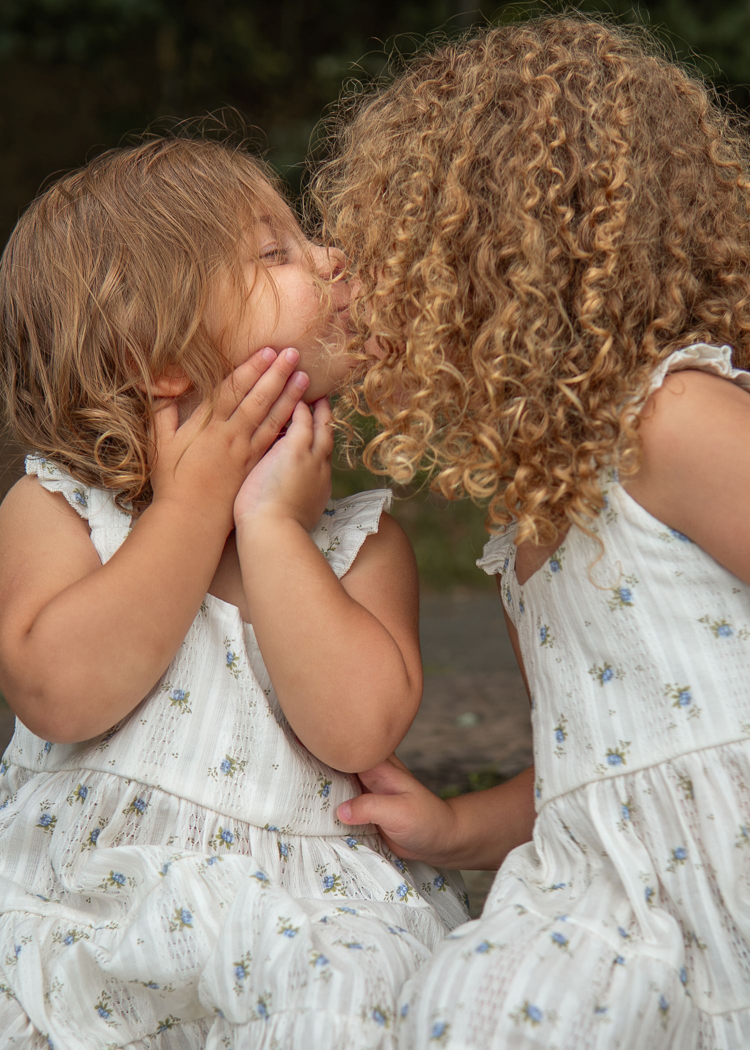 Two young girls in matching floral white dresses sharing a sweet kiss outdoors.