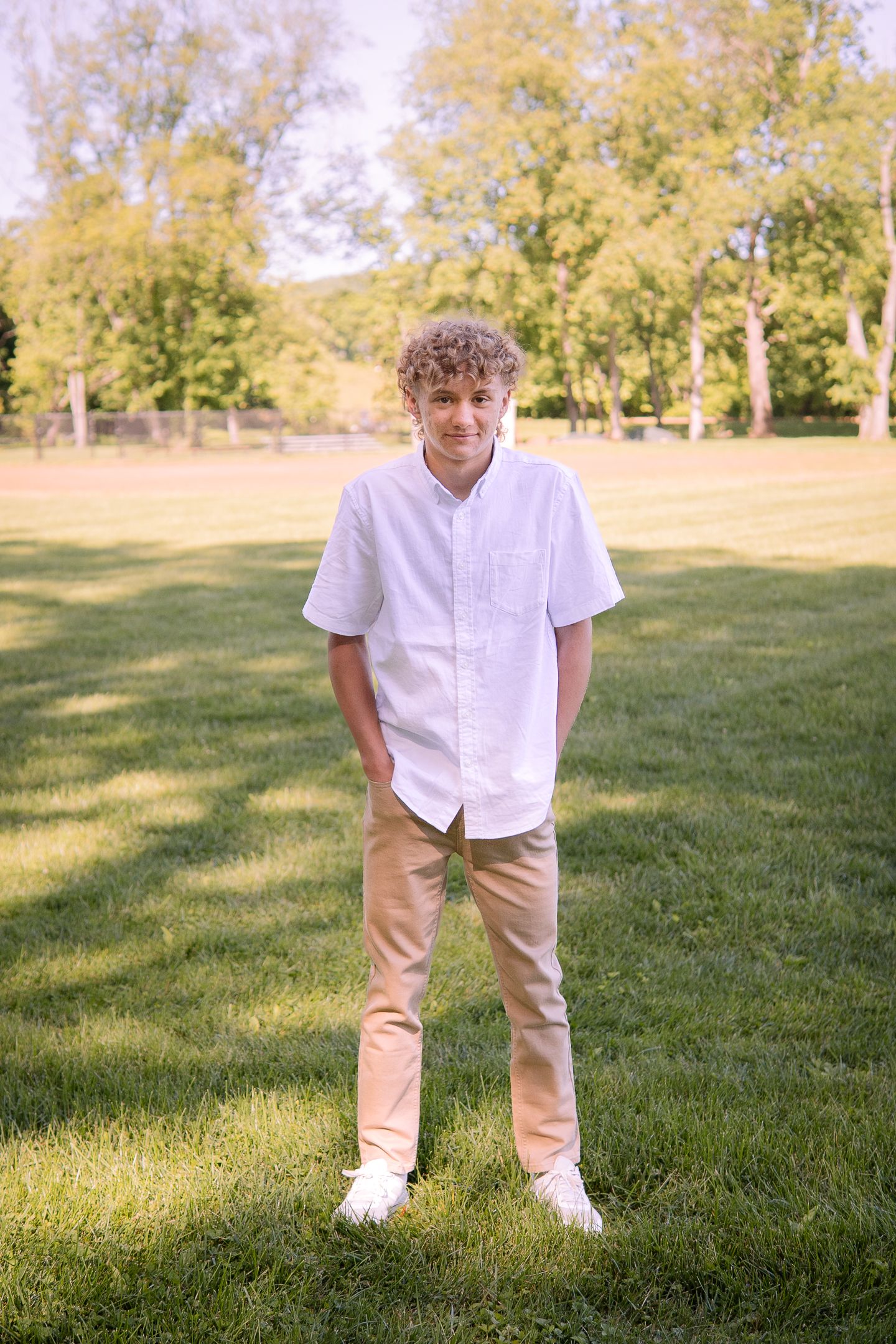 A casual portrait of someone in white shirt and khaki pants standing on grass with trees in background.
