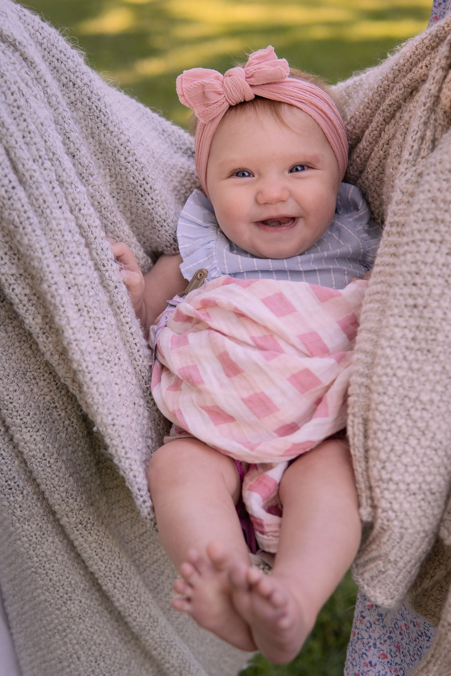 A happy baby wearing pink bow headband and checkered outfit lies in a hammock outdoors.