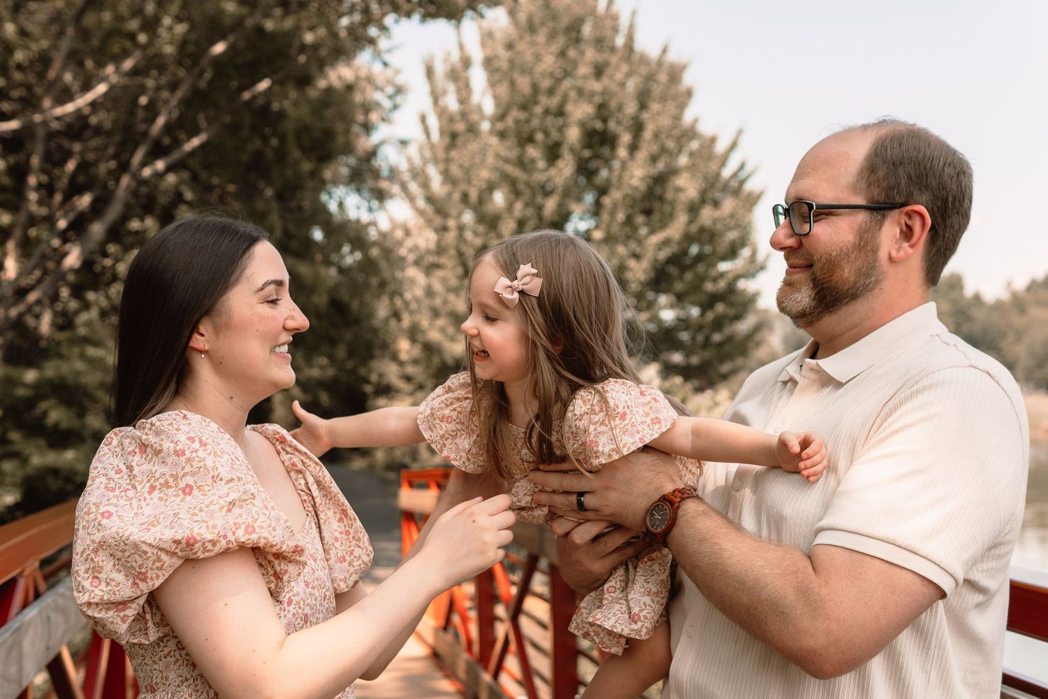 Happy family portrait with parents and young daughter on outdoor bridge surrounded by trees.