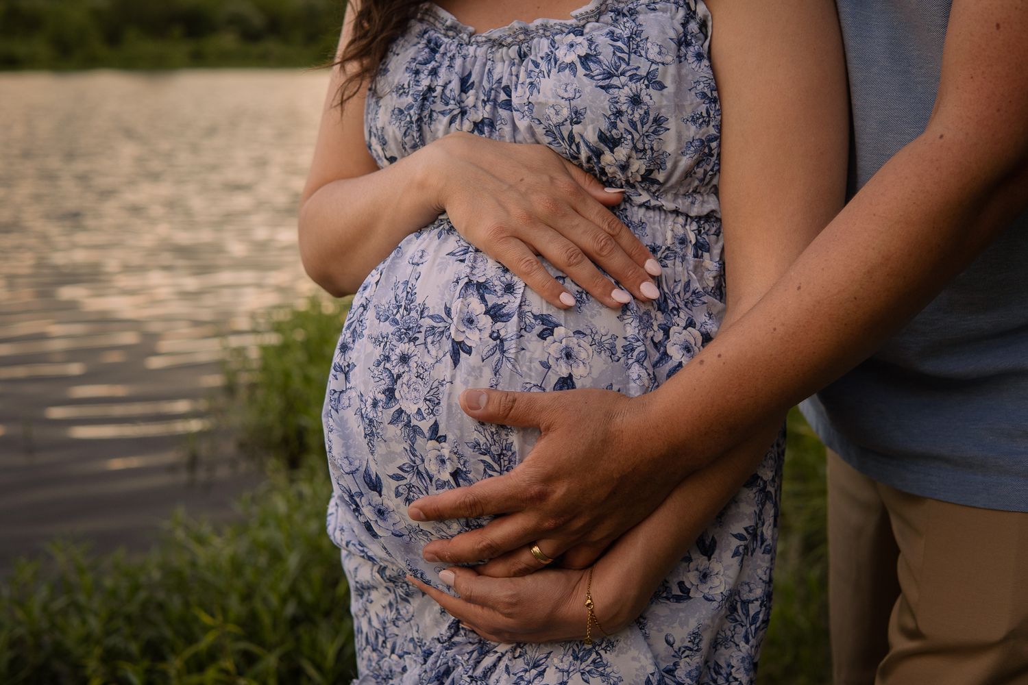 Close-up of couple cradling pregnant belly near lake, showing wedding ring.