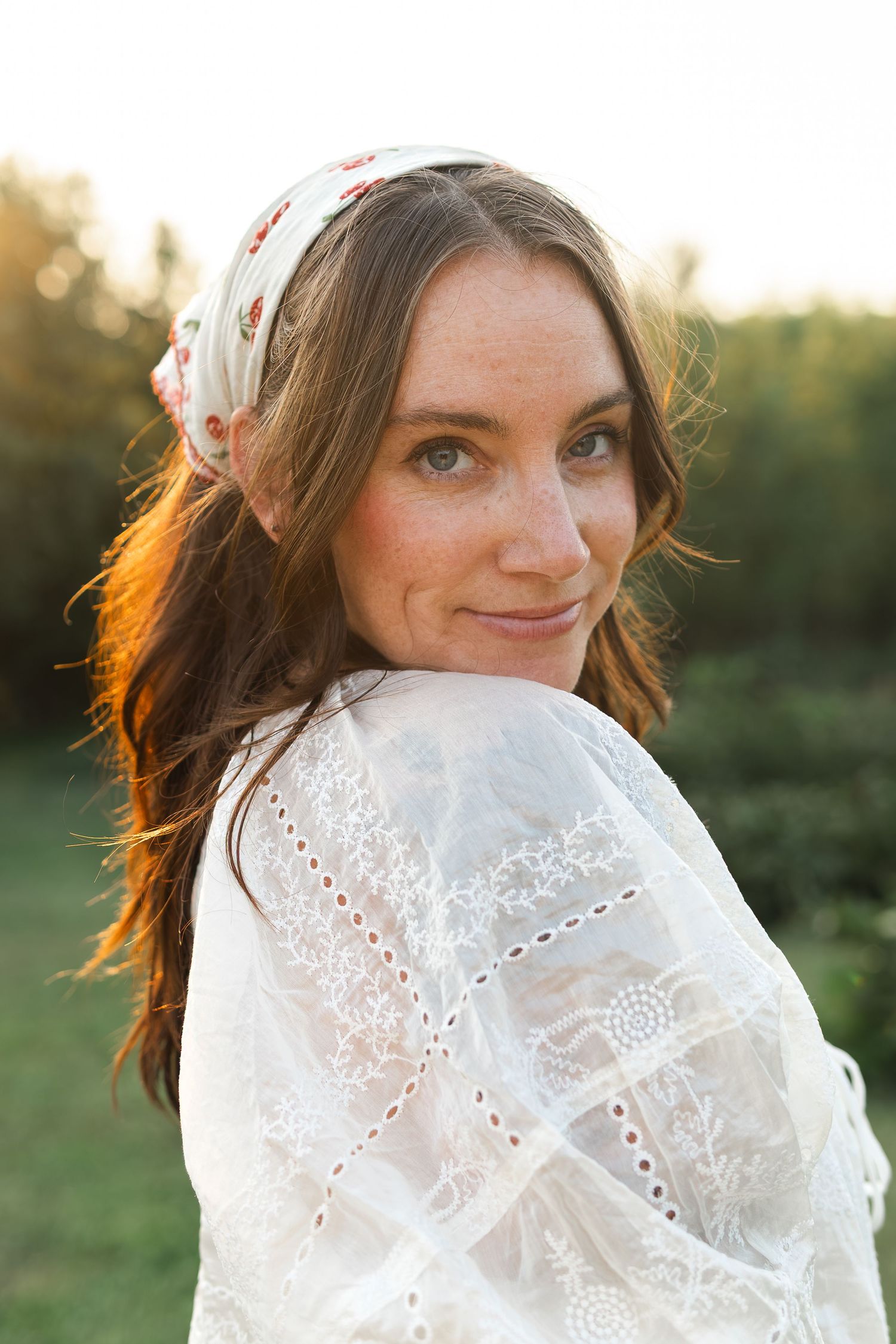 Young woman with freckles wearing white lace dress and floral headscarf smiling outdoors at golden hour.