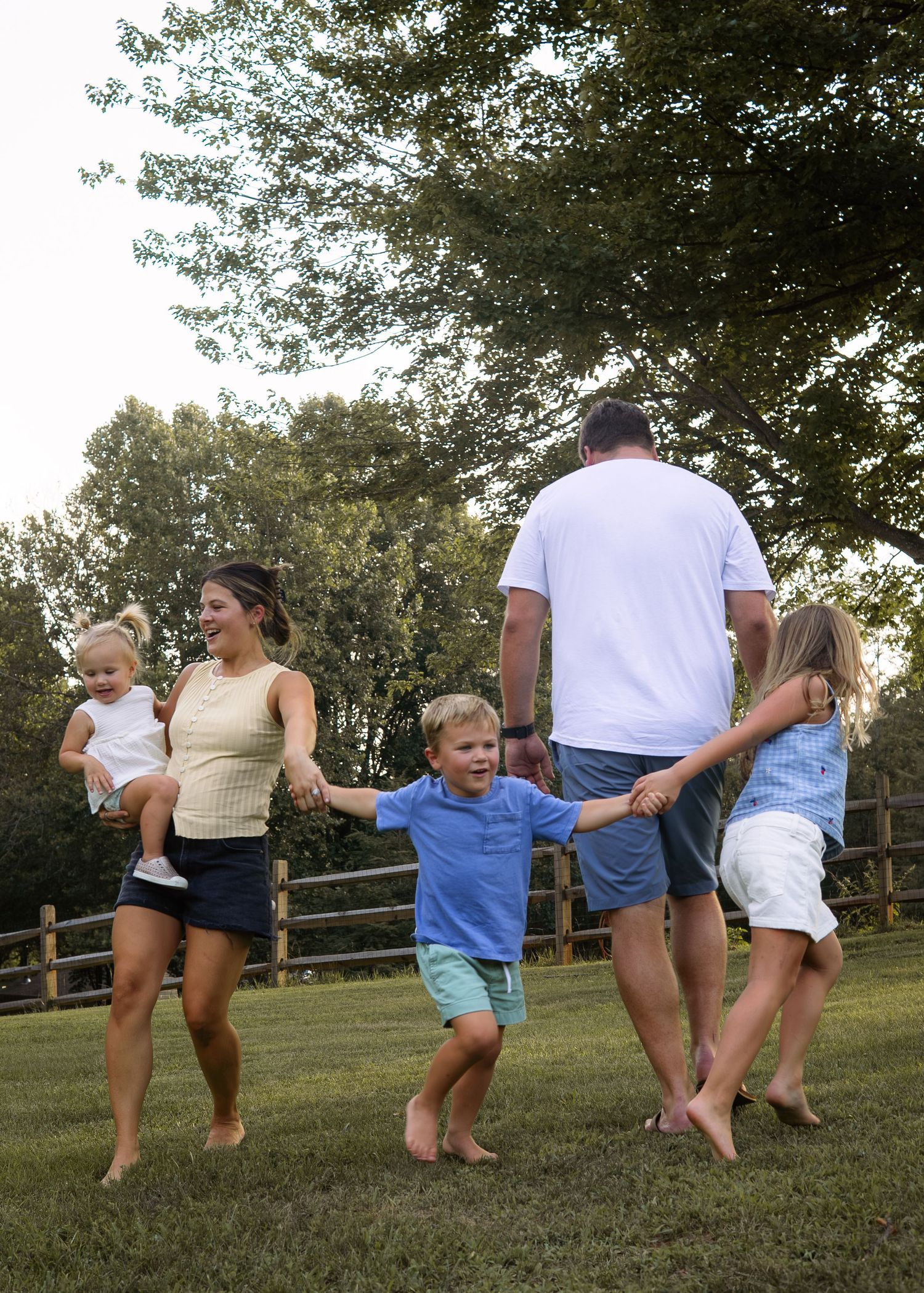 A happy barefoot family of five holds hands while walking together across a sunny green yard.