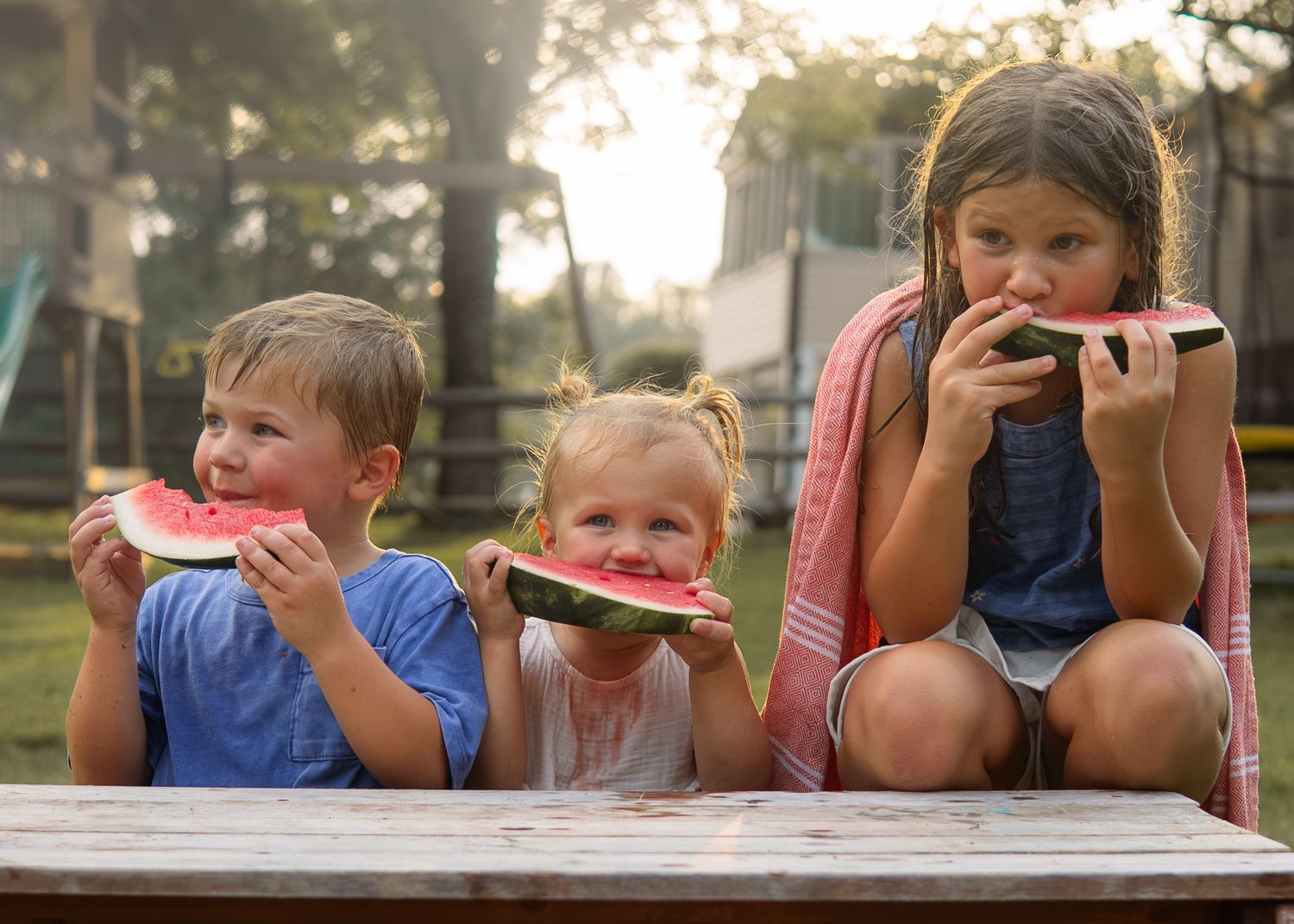 Three young children eating fresh watermelon slices at an outdoor table.