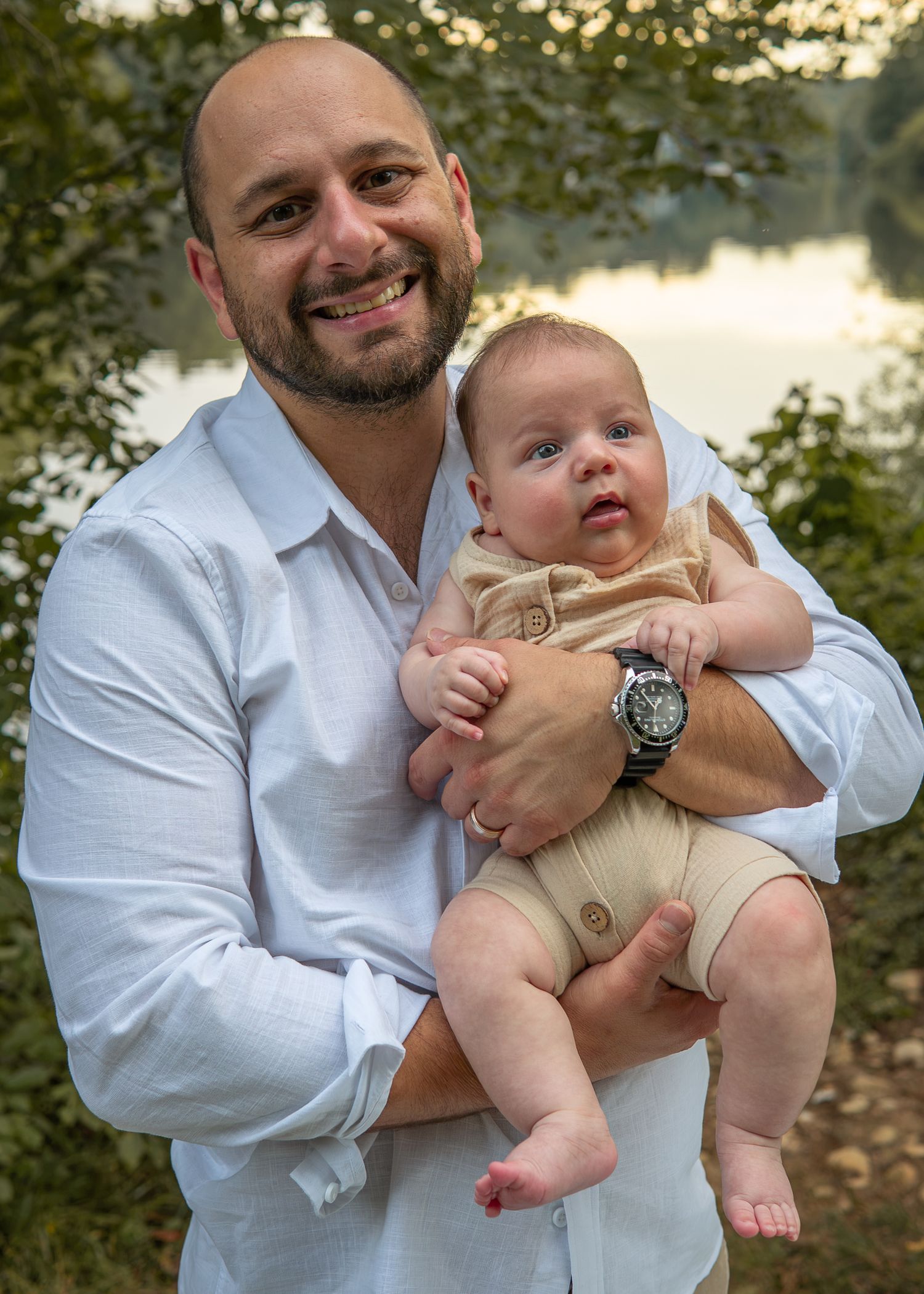 Smiling bald father in white shirt holding his infant baby boy outdoors near a lake.