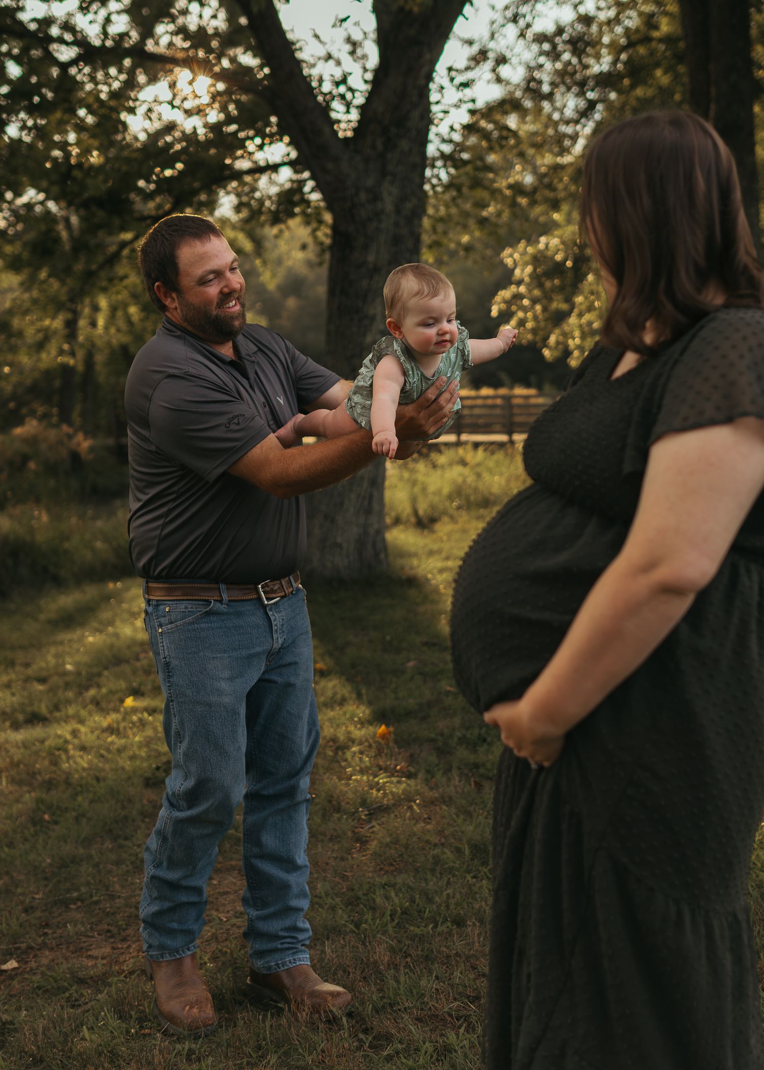 A man holds a baby outdoors at golden hour while a pregnant woman in a black dress watches in a lush green setting.