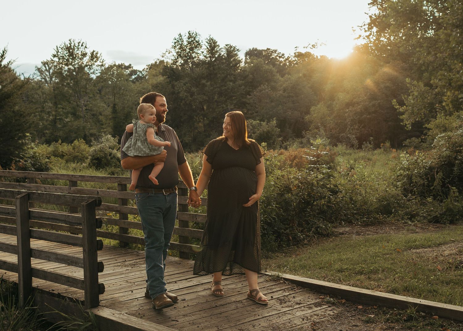Young family holds hands while strolling across wooden bridge in lush green park at sunset.