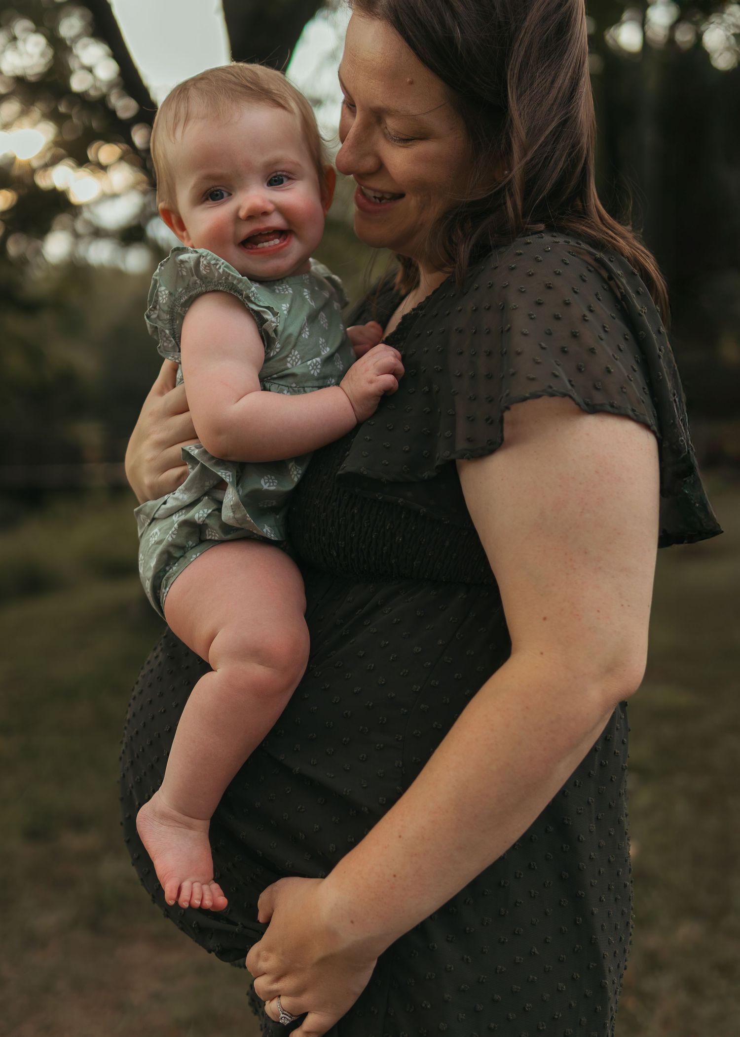 A smiling baby in a green romper being held by a woman in a dark dress outdoors.