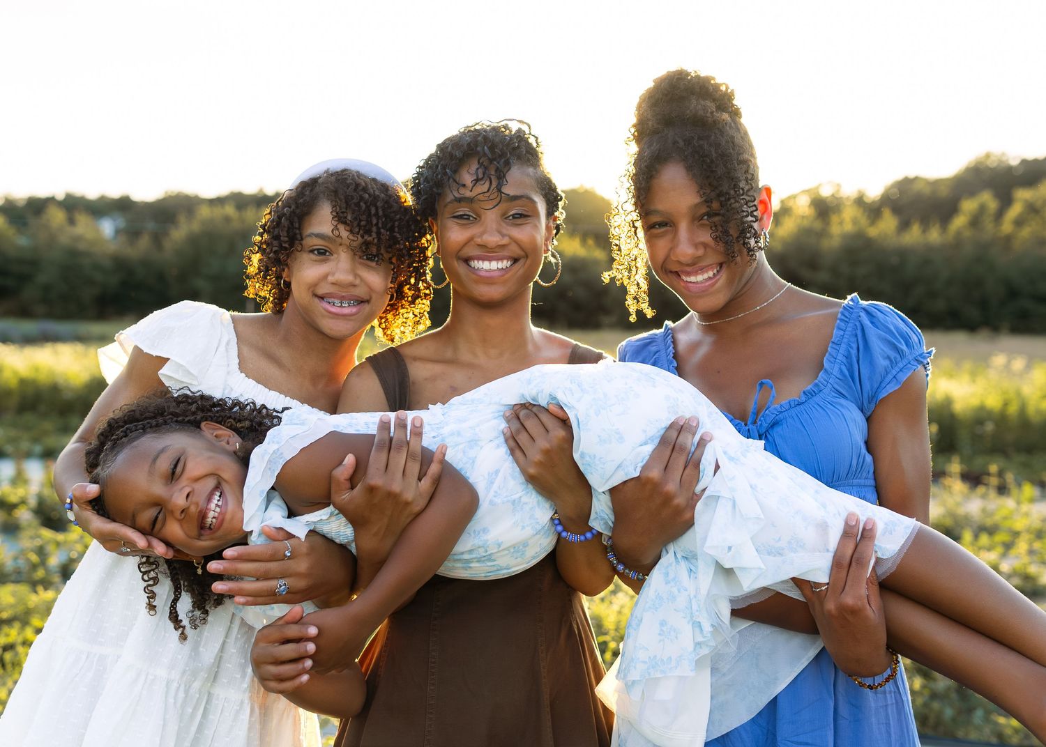 A smiling mother holds a baby while surrounded by three daughters in a sunny outdoor field setting.