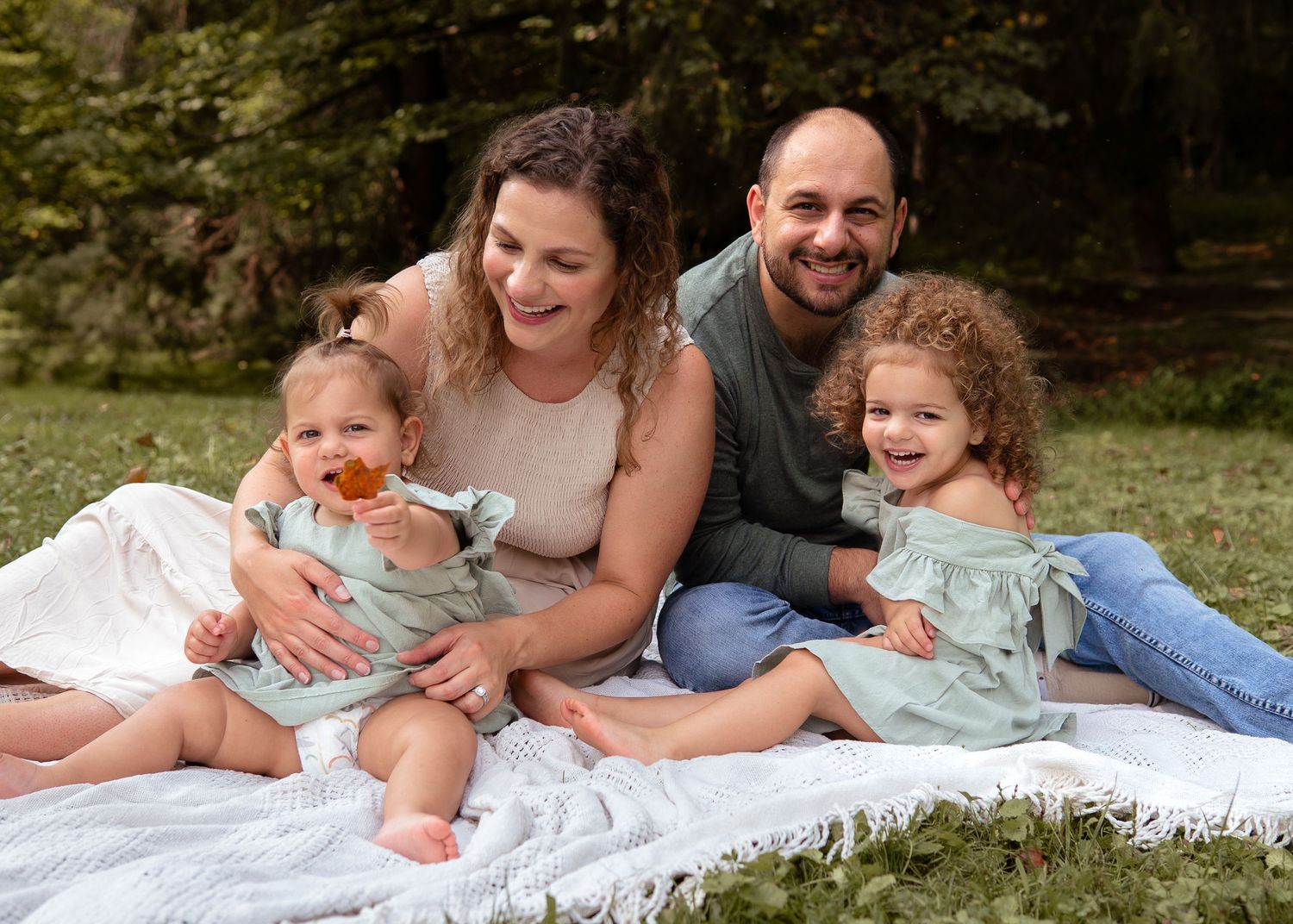 Happy family of four sitting on white blanket outdoors with two young daughters smiling.