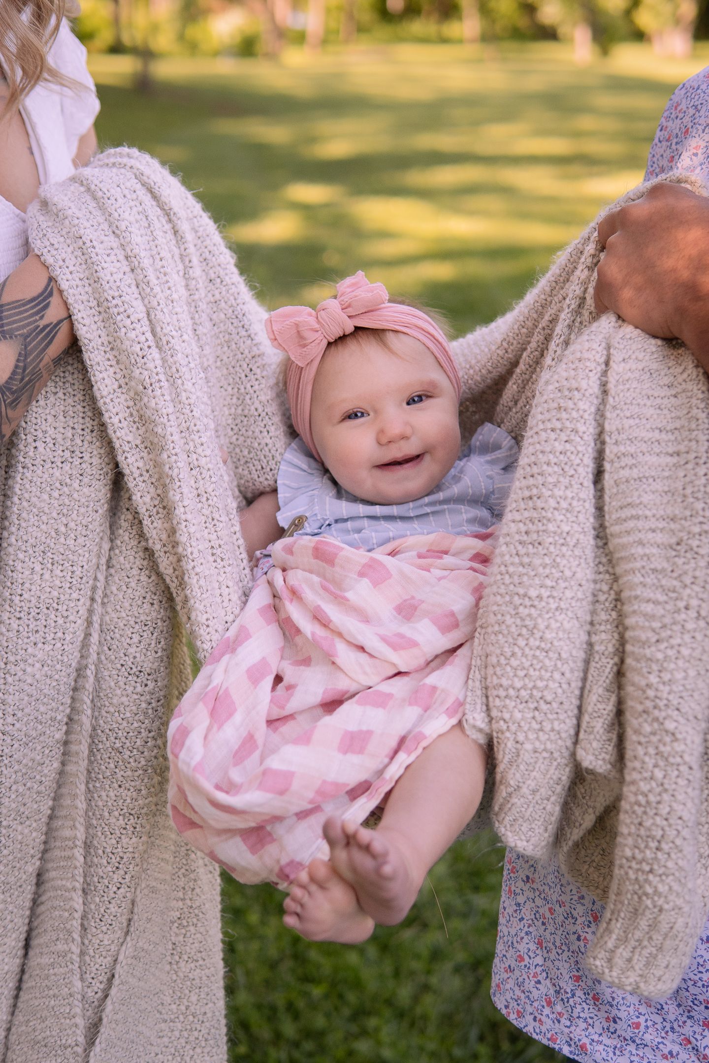 A smiling baby wearing pink being held in a knitted blanket outdoors on a sunny day.