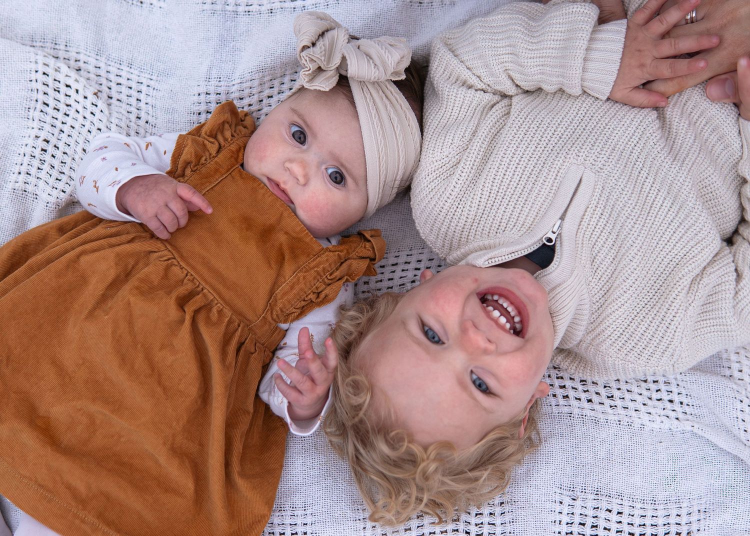 Two babies lying on a white lace blanket, one in an orange dress and one in a striped outfit, smiling.