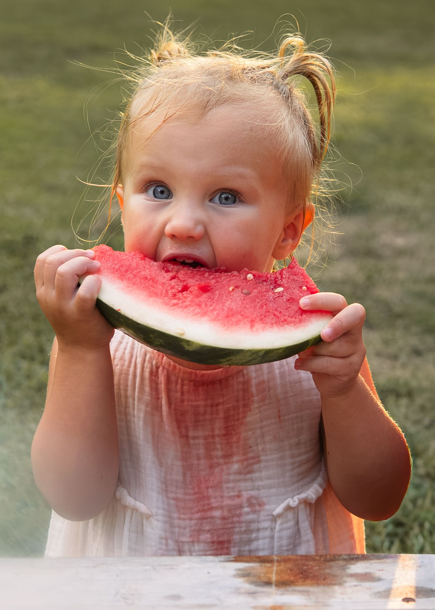 Cute toddler girl with blue eyes eating a large watermelon slice outdoors.