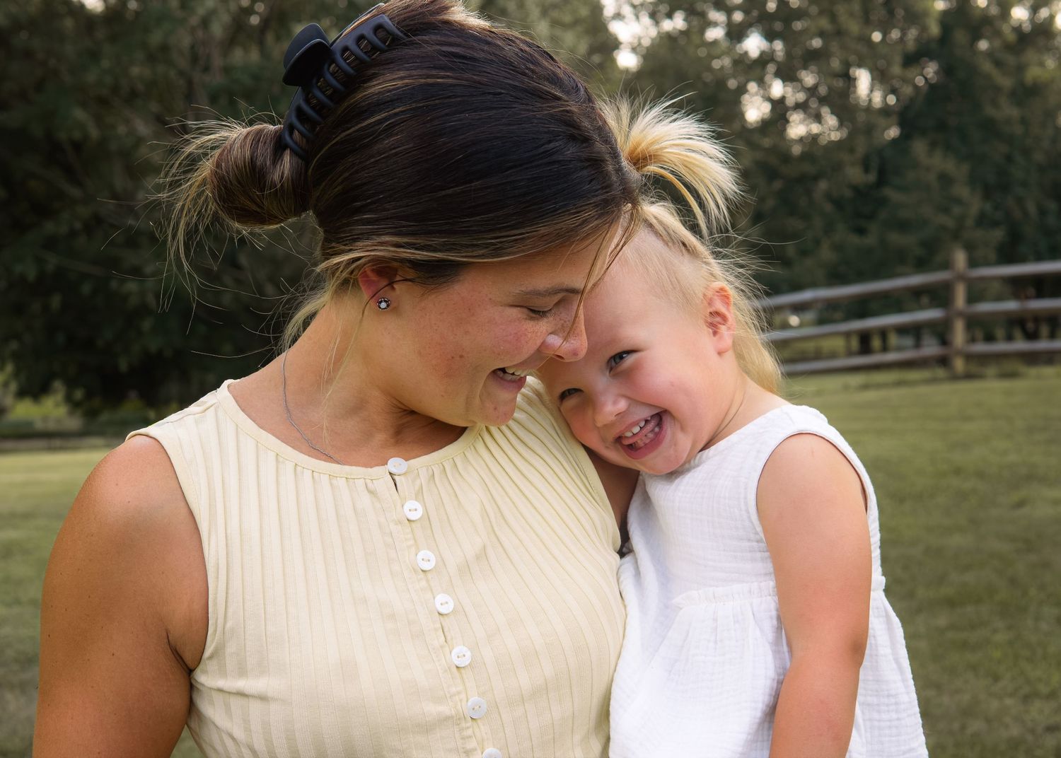 A smiling mother and laughing toddler girl share a joyful moment together outdoors on a green lawn.