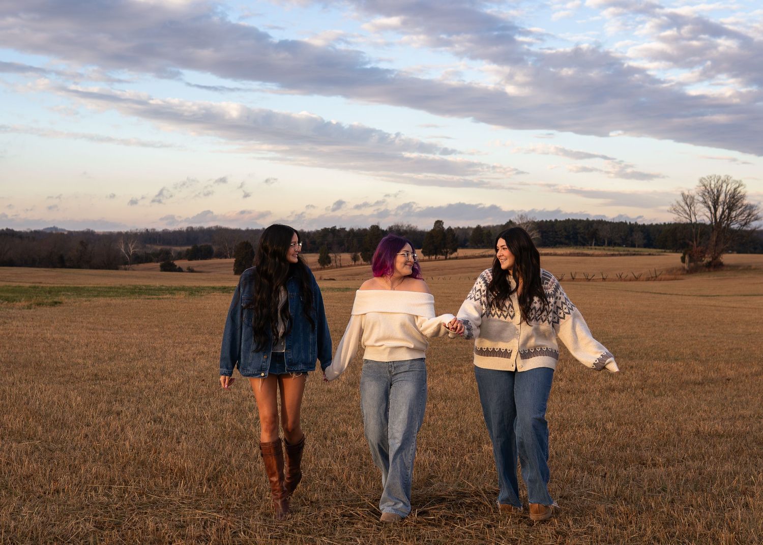 Three young women walk hand-in-hand through a golden field at sunset under a dramatic cloudy sky.