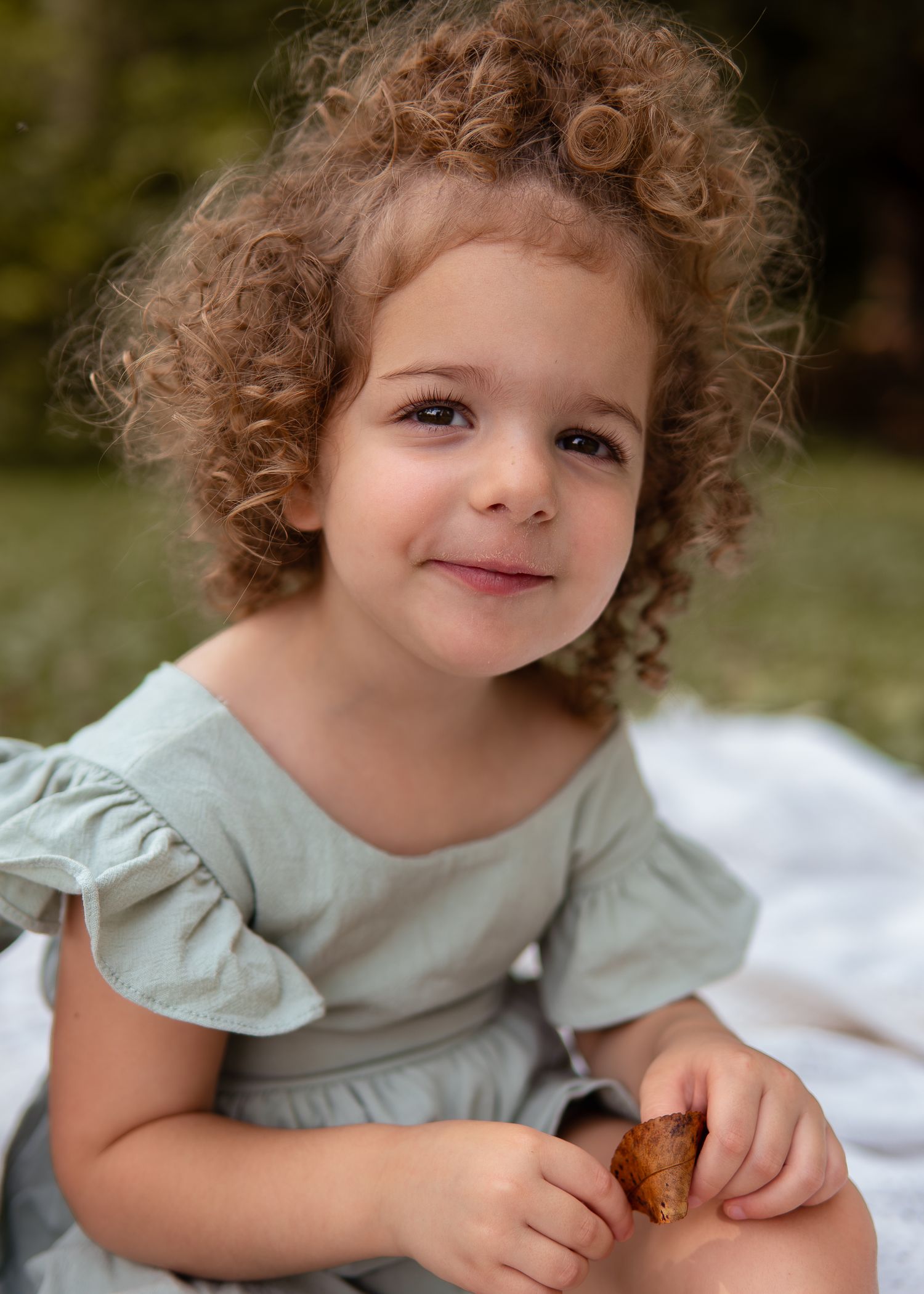 Adorable curly-haired toddler girl in sage dress smiling outdoors on a blanket.