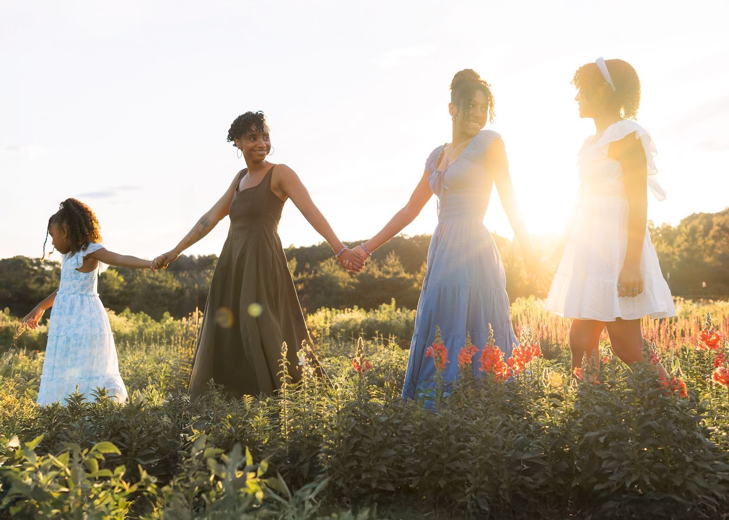 Four women and girls hold hands in a sunlit field wearing colorful dresses during a golden hour outdoor photo session.