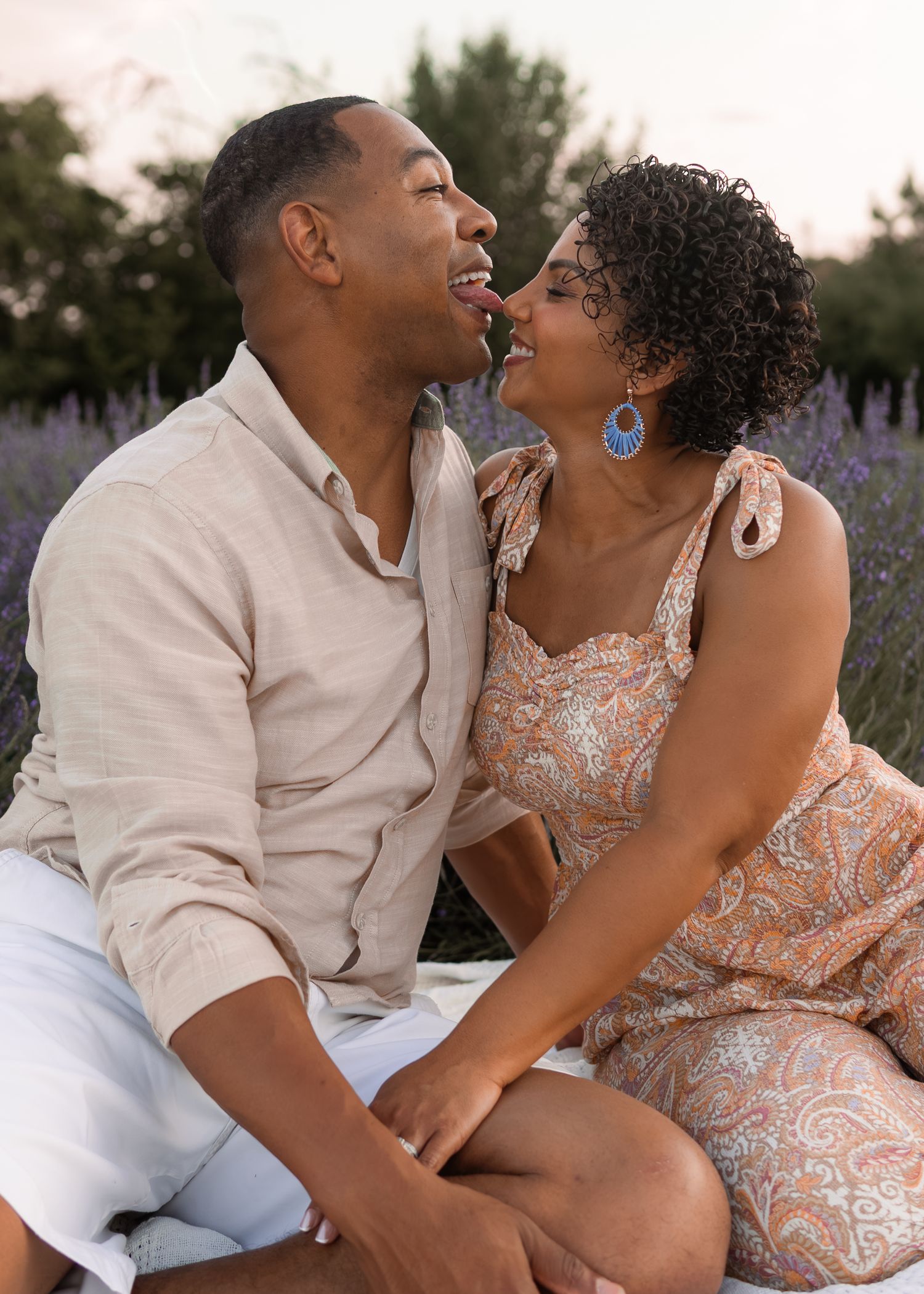 A happy couple sits closely together in a lavender field, sharing a romantic moment and smiling at each other.