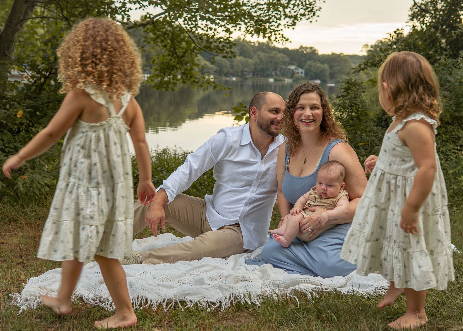 Family of five poses on a blanket by a river at sunset, parents sitting with newborn while two young girls play nearby.