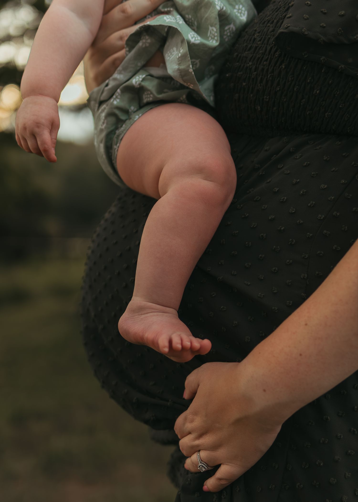 Close-up of adorable baby legs and feet being held by pregnant mother in black dress outdoors.
