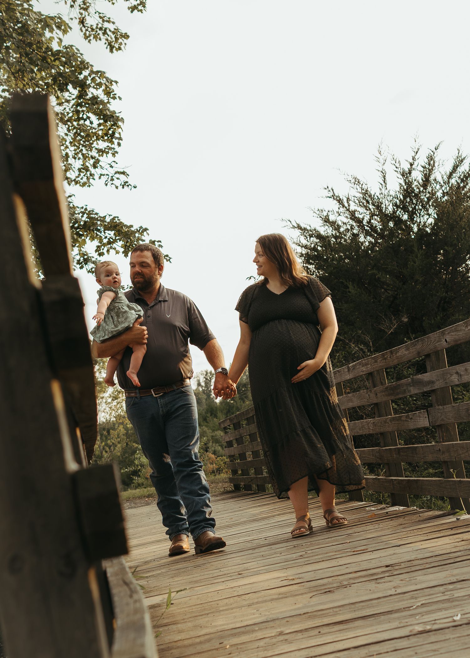 Pregnant woman in black dress holds hands with man carrying toddler on wooden bridge outdoors.
