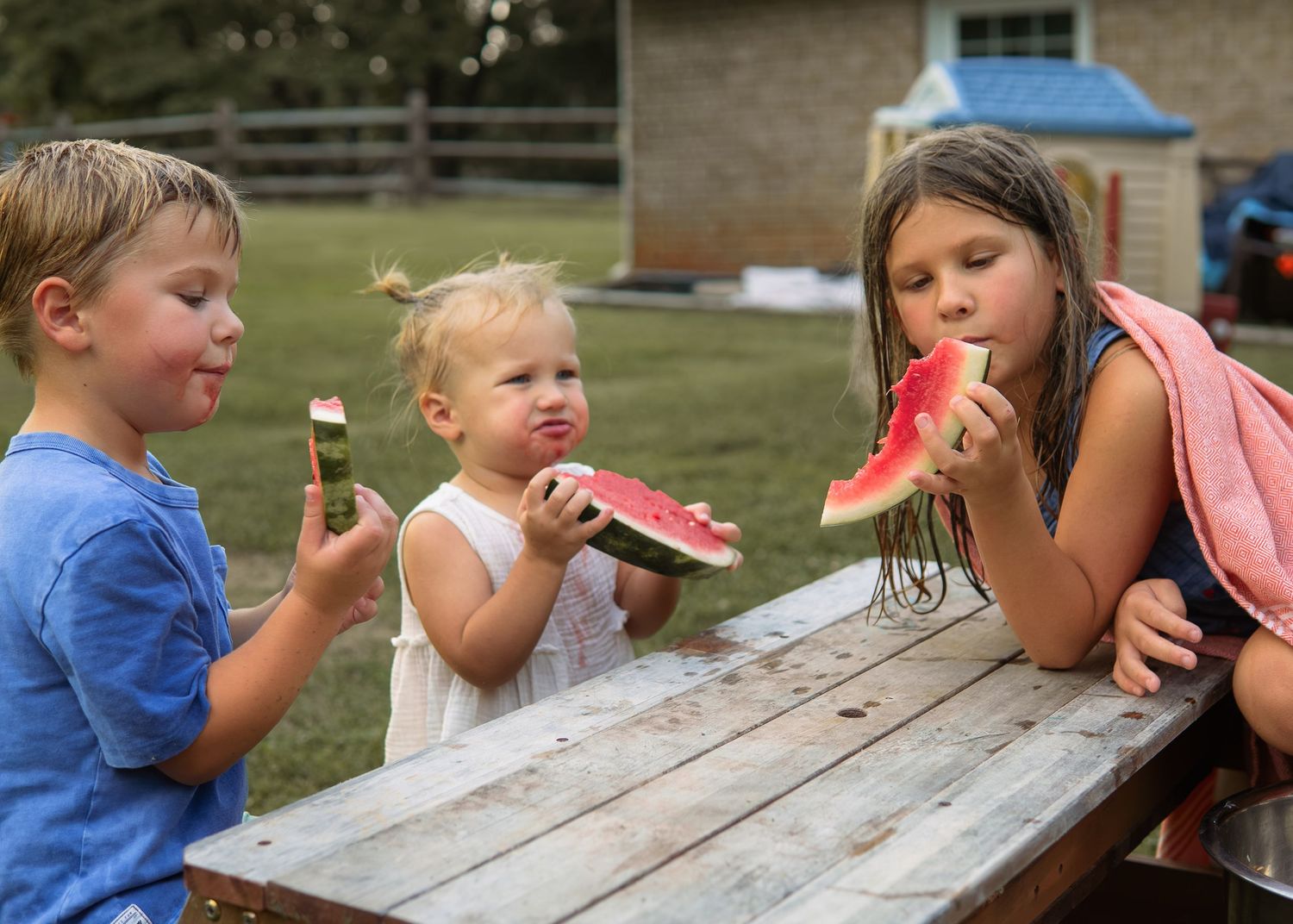 Three young children eating fresh watermelon slices at a wooden picnic table outdoors on a farm.