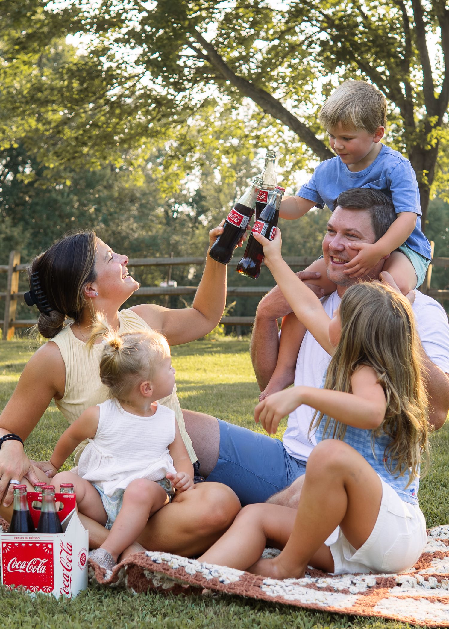 Family of five enjoying a Coca-Cola picnic outdoors, toasting bottles together on a sunny day in a lush green park.