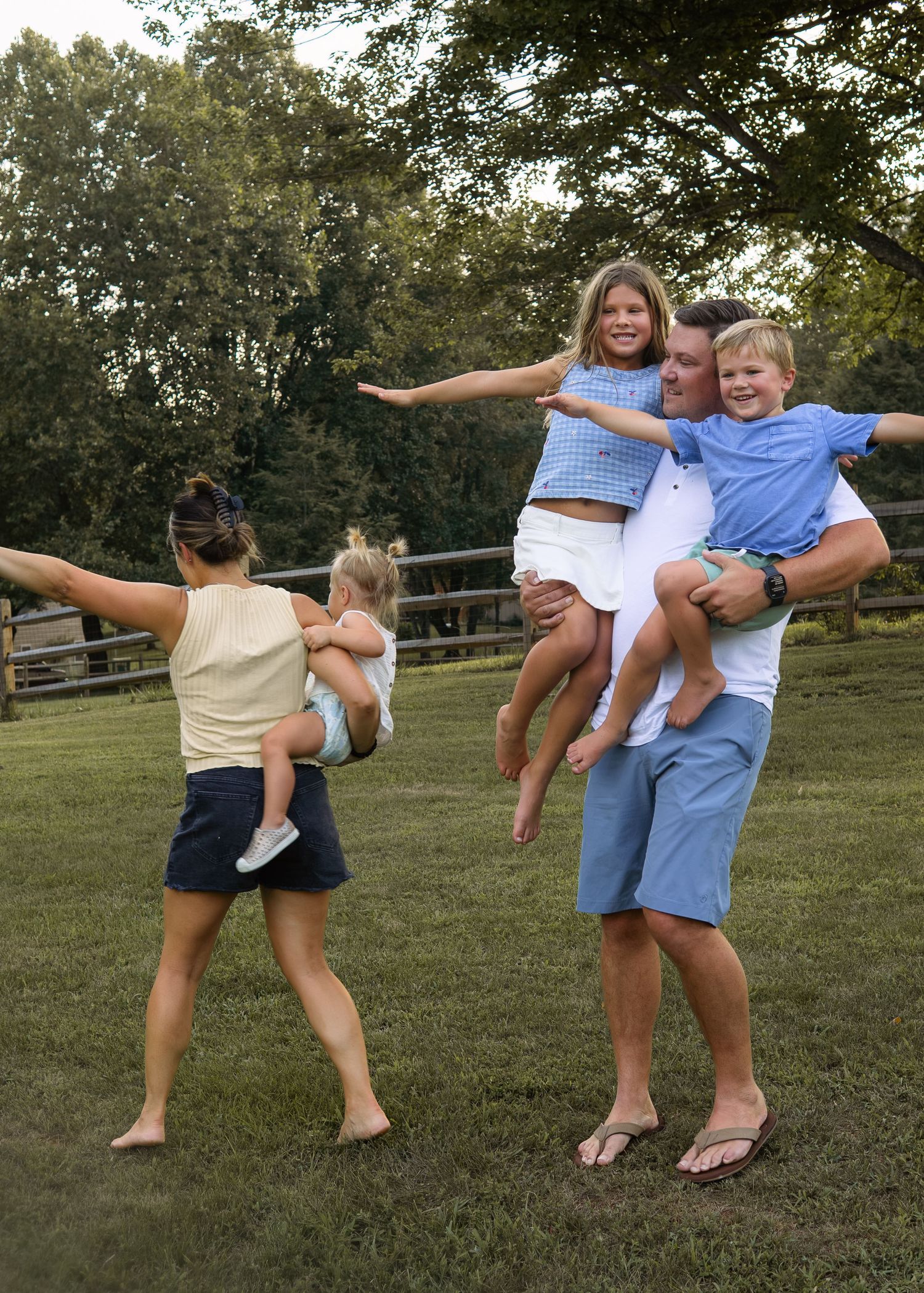 A playful family with two parents holding three children with arms outstretched in a lush green outdoor setting.