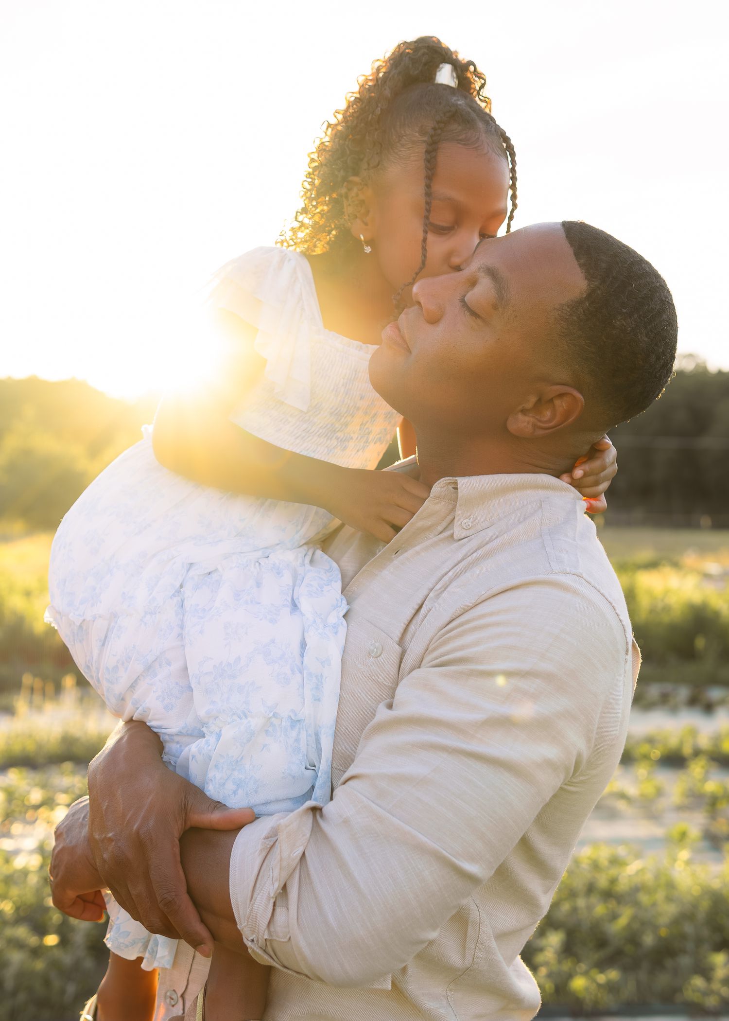 A father holds his young daughter as she kisses his cheek during a golden sunset outdoor photo session.