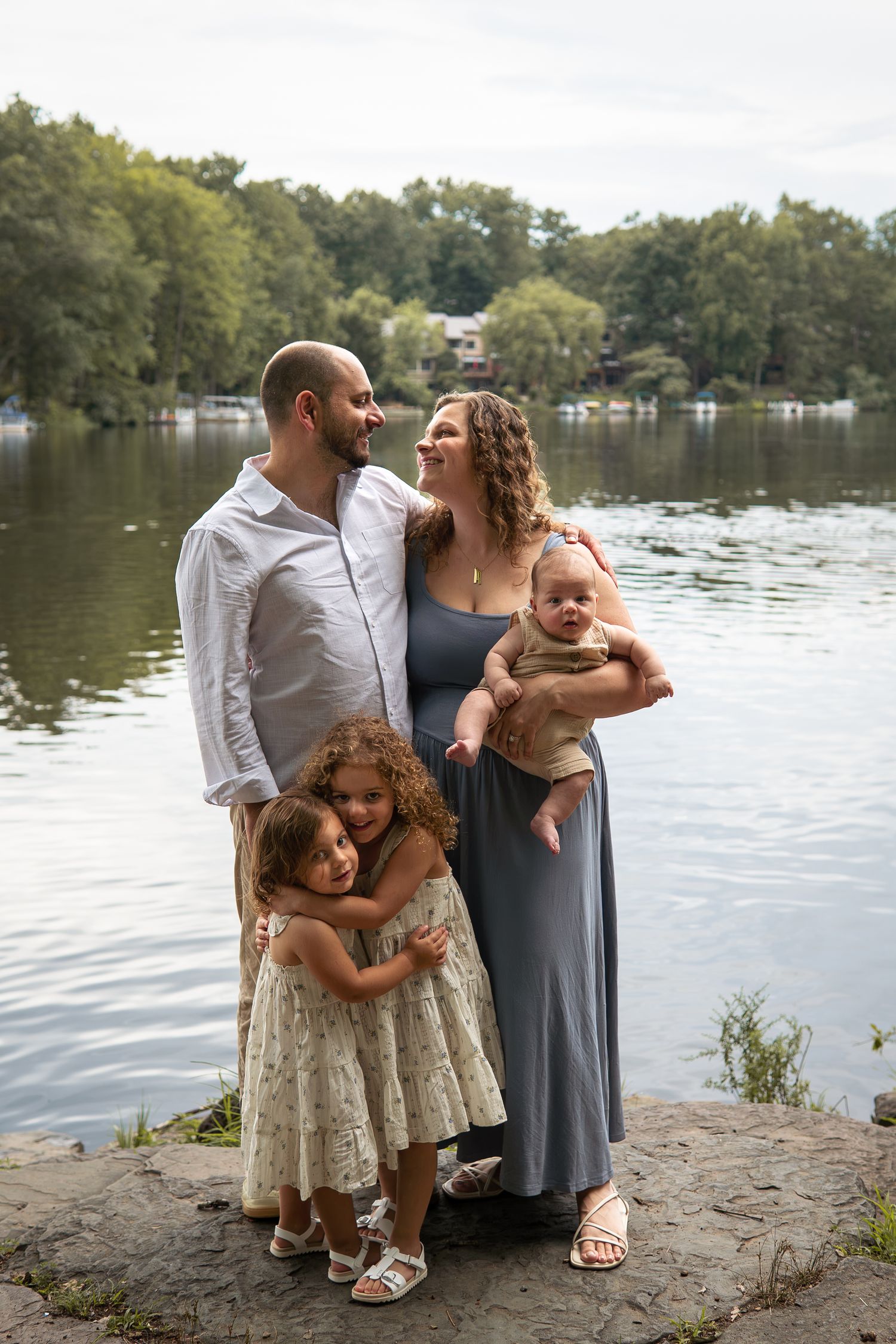 Family of five posing by a lake, parents smiling with baby and two young girls hugging in front.