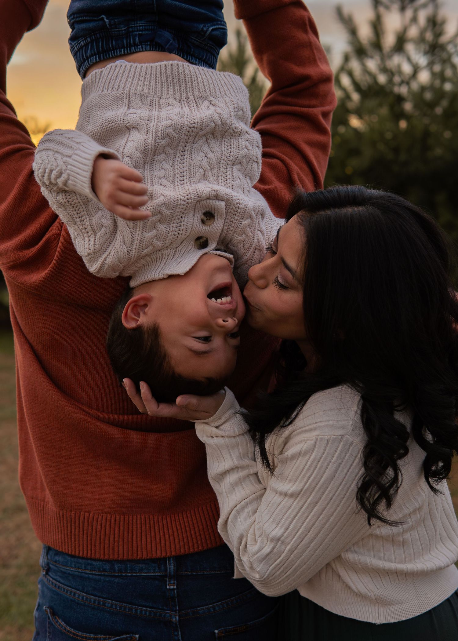 Parents playfully kissing their laughing toddler held upside down at golden hour outdoors.