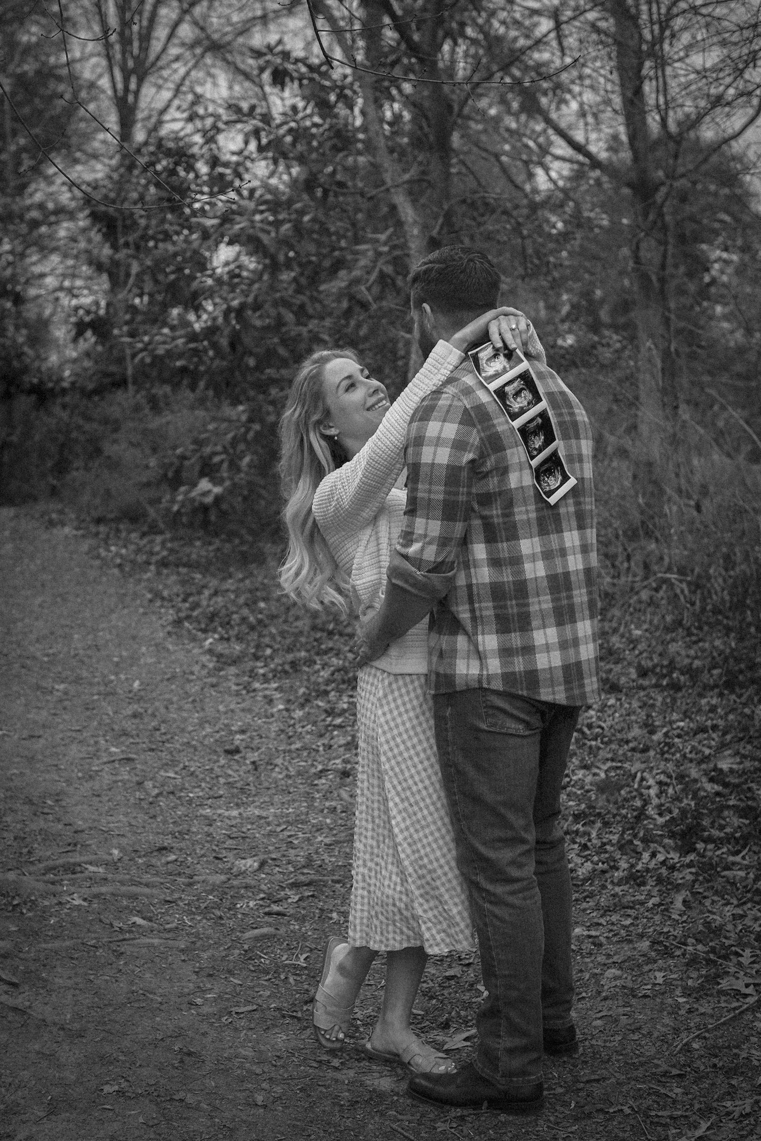 A couple embraces on a wooded path in black and white, with the woman wearing a patterned dress and man in plaid.