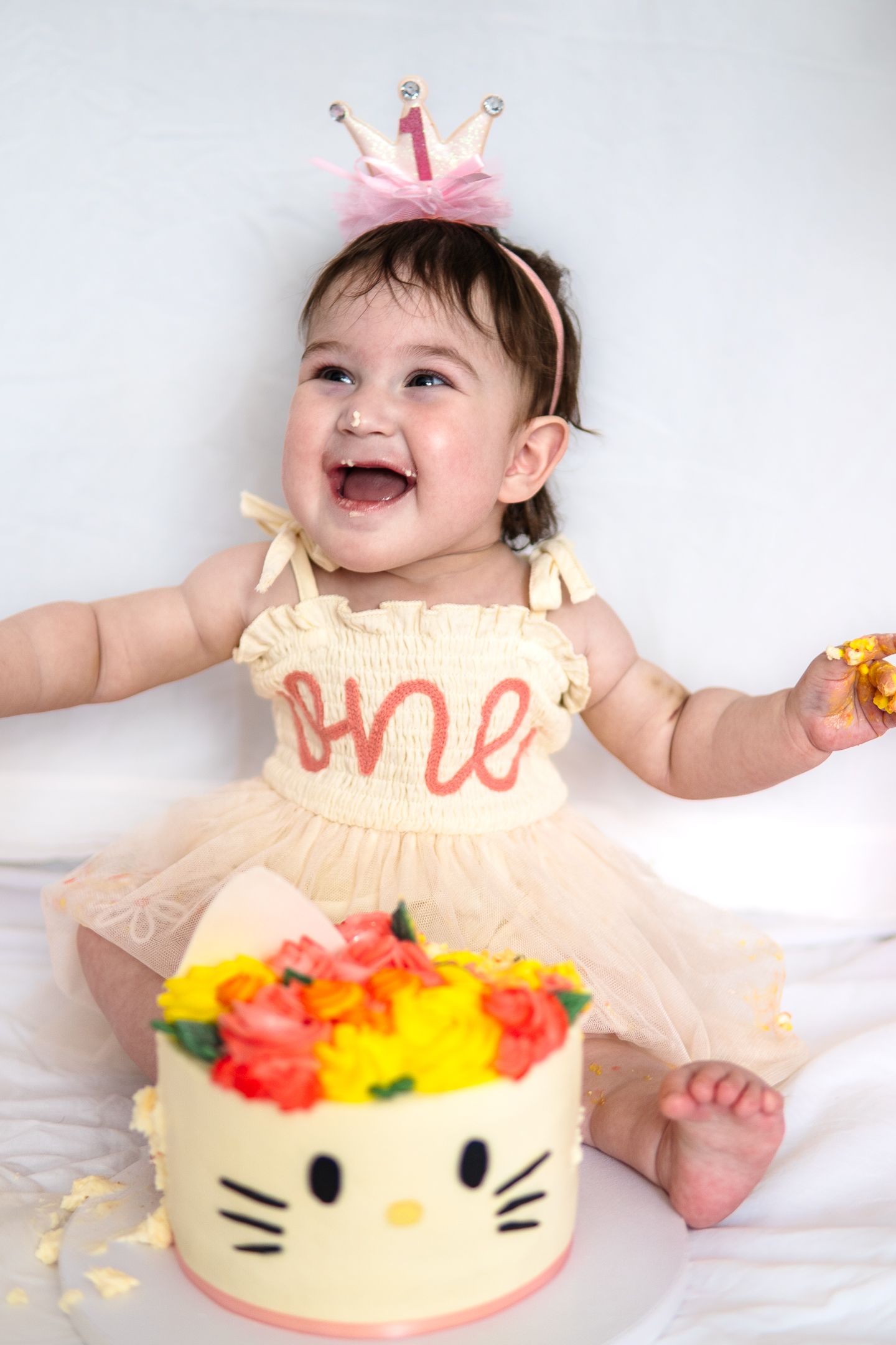 Joyful baby wearing crown headband and cream dress laughs while celebrating first birthday with Hello Kitty cake.
