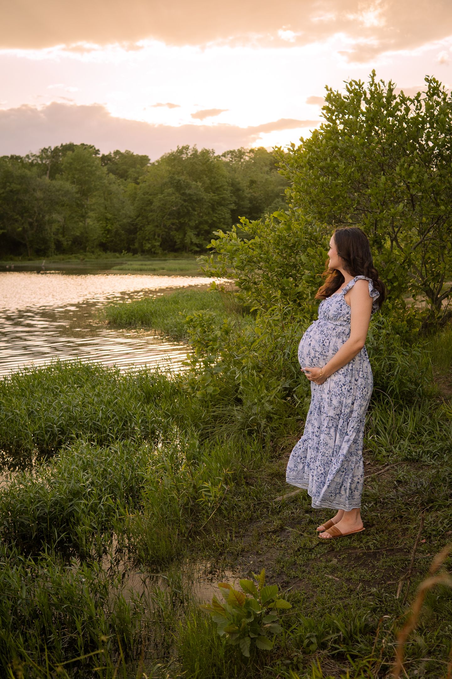 A maternity photo of an expectant mother in a floral dress standing by a lake at sunset.