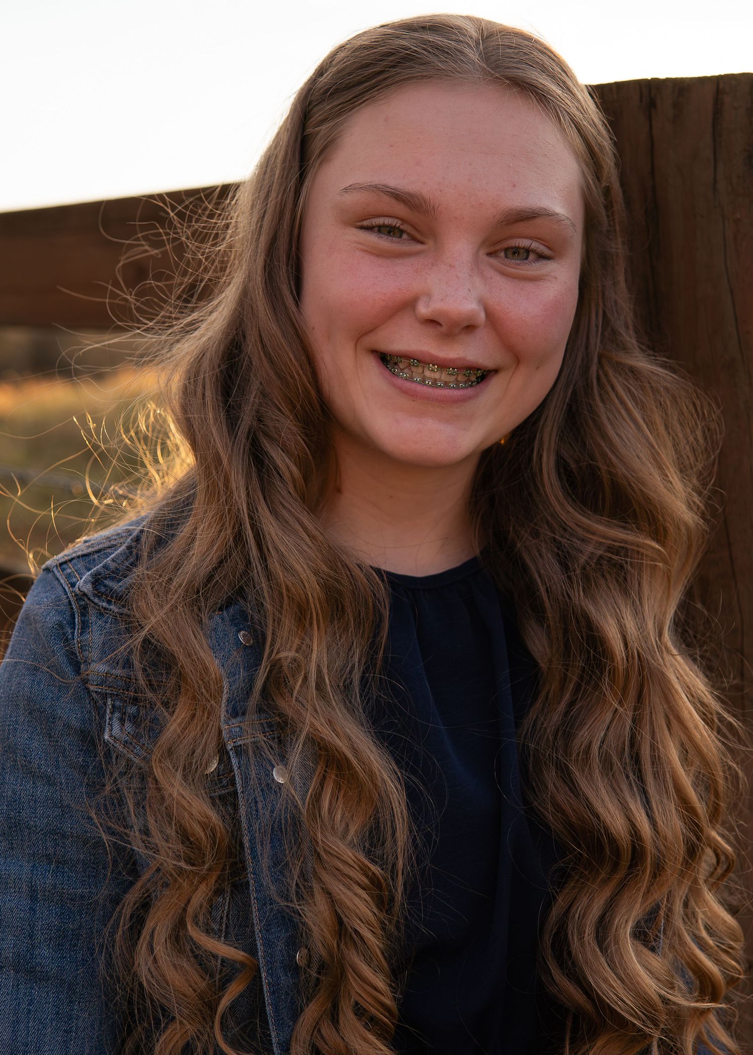 Smiling teenage girl with braces and long wavy hair in denim jacket at sunset.