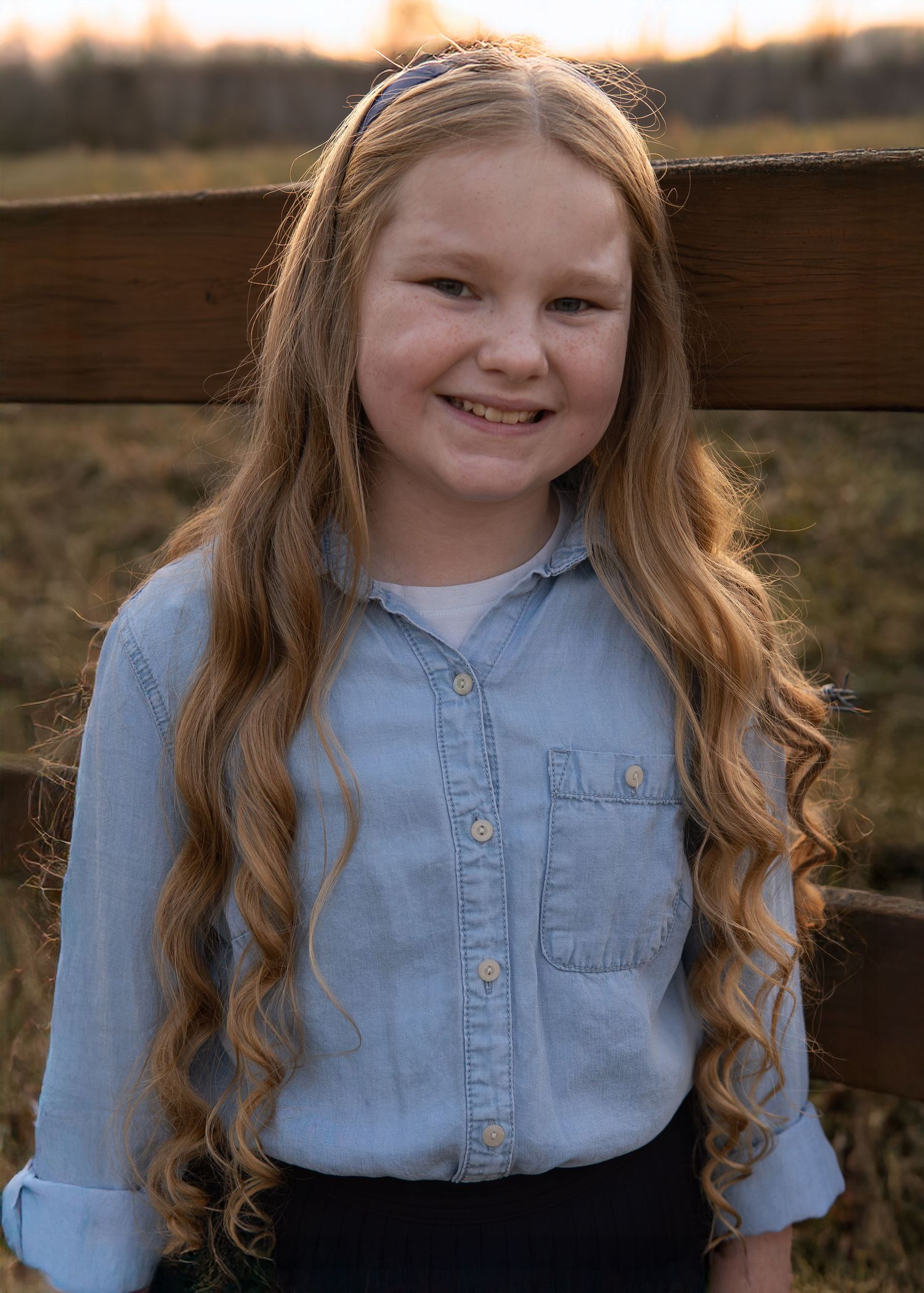 Smiling girl with long curly red hair wearing a light blue denim shirt, leaning against a wooden fence outdoors.