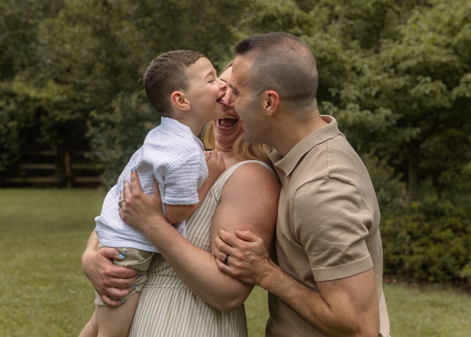 A joyful family moment with parents laughing and holding their young son outdoors in a lush green park.